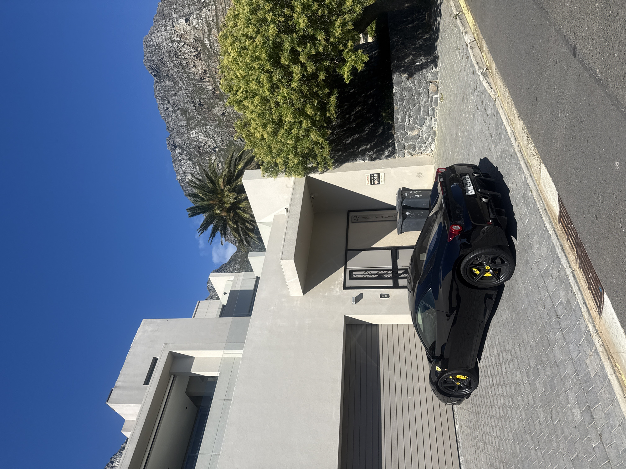 The image depicts a black sports car parked on a paved driveway next to a modern, multi-story building with white walls and minimalist design. The driveway abuts a grey asphalt road. Behind the building, Table Mountain rises prominently under a clear blue sky, with a palm tree and other greenery visible in front of the mountain. The scene appears to be a residential area in Cape Town, South Africa.