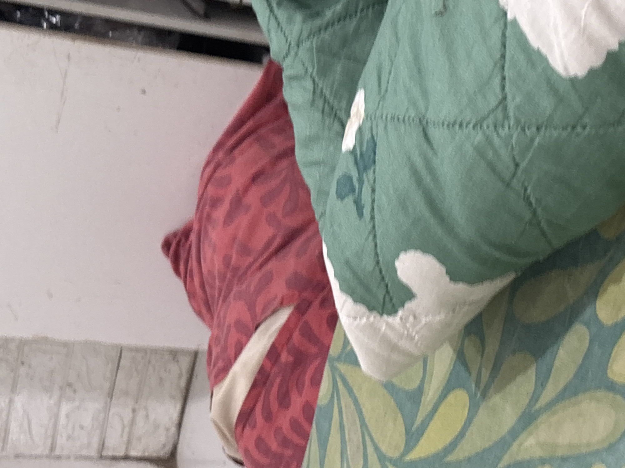 The image shows a close-up view of a bed with multiple layers of bedding. The top layer consists of a red blanket with a patterned design. Below that, a teal and light green patterned sheet is visible. The third visible layer is a green comforter with a quilted diamond pattern and white decorative stitching. The scene appears to be indoors, likely in a bedroom. The lighting is soft and diffused. There are no visible people in the image. The overall impression is one of comfort and domesticity.
The location in the city of North Jakarta, Indonesia, is not determinable from the image content.