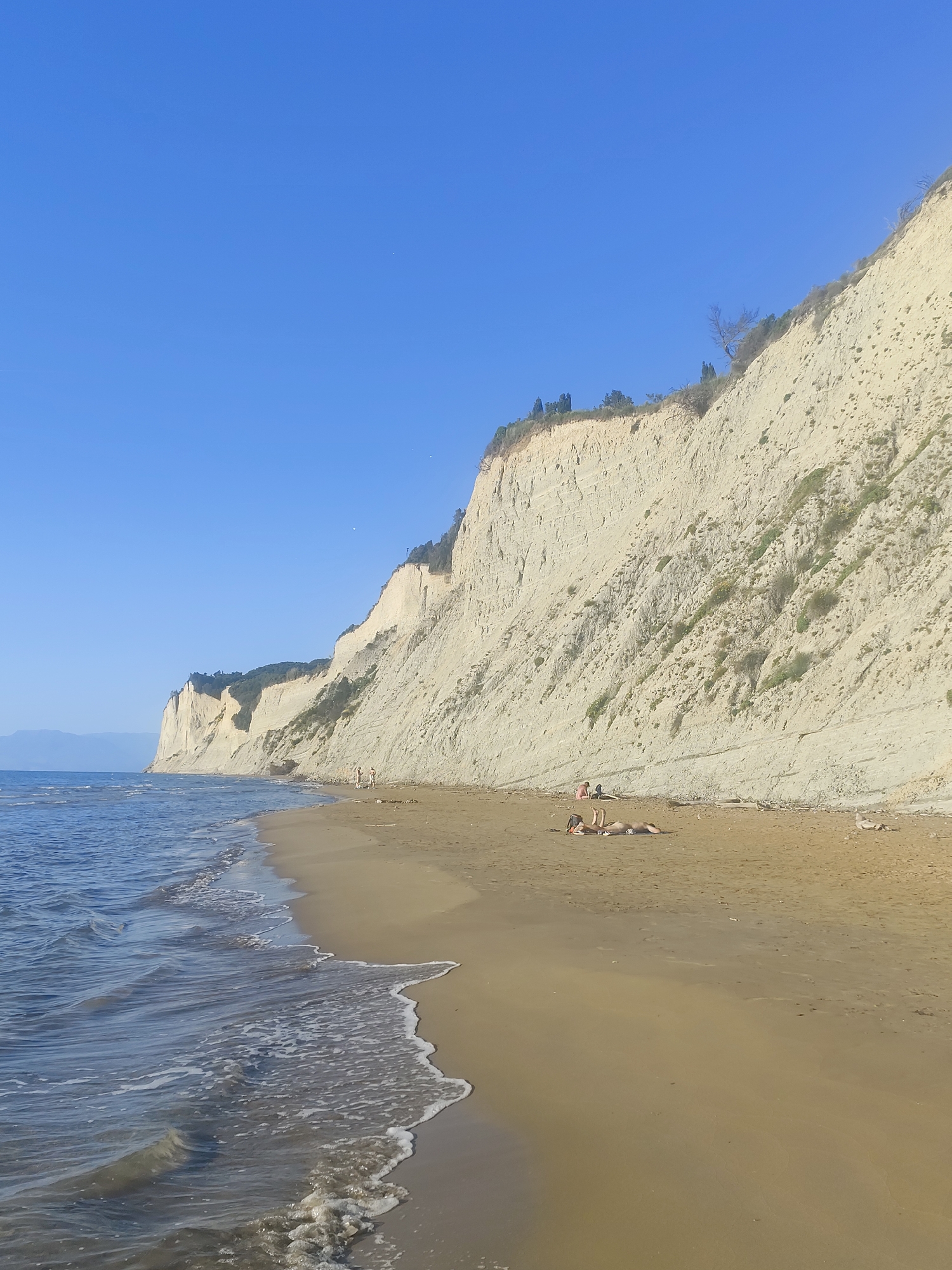 The image depicts a beach scene. In the foreground, waves are washing onto a sandy shore. The beach extends towards the right, where several people are lying down, seemingly sunbathing. In the background, a tall, light-colored cliff rises vertically from the beach. Some trees and vegetation grow on top of the cliff. The sky is clear and blue. The location is in the city of Unknown, Unknown.