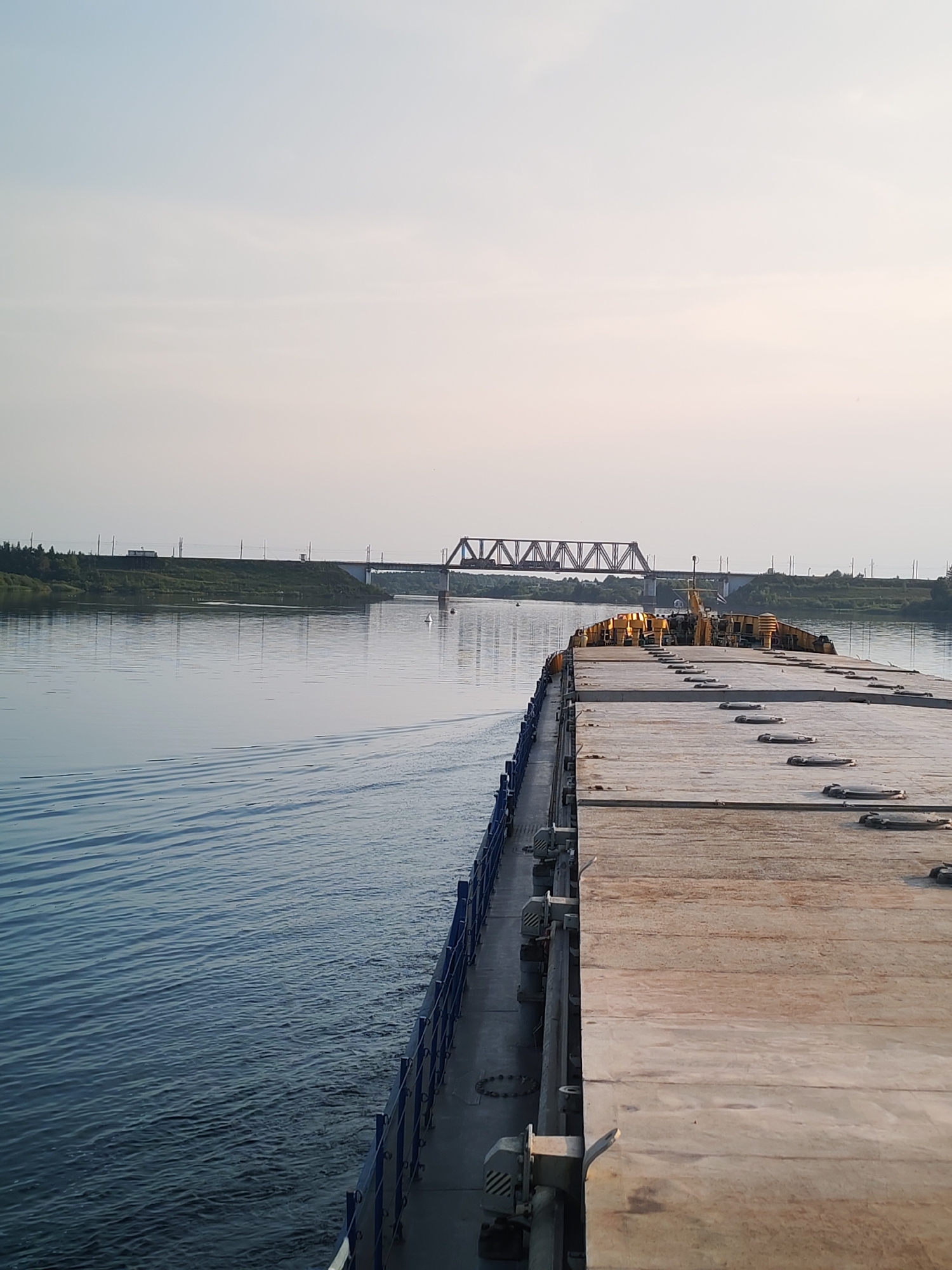 The image shows a view from the deck of a barge on a calm river. The barge's flat deck, made of large, weathered wooden planks, occupies the right side of the frame, with a blue railing visible along its edge. The water to the left of the barge is a deep blue, showing gentle ripples from the barge's movement. In the distance, the river widens, and a green, tree-lined bank is visible. A railway bridge with a distinctive truss structure spans the river in the mid-ground. On the far bank, there are signs of human activity, including what appear to be railway tracks and a small structure. The sky is a pale, hazy blue, suggesting either early morning or late evening light. The overall scene is peaceful and industrial, evoking a sense of transportation and transit along a waterway.