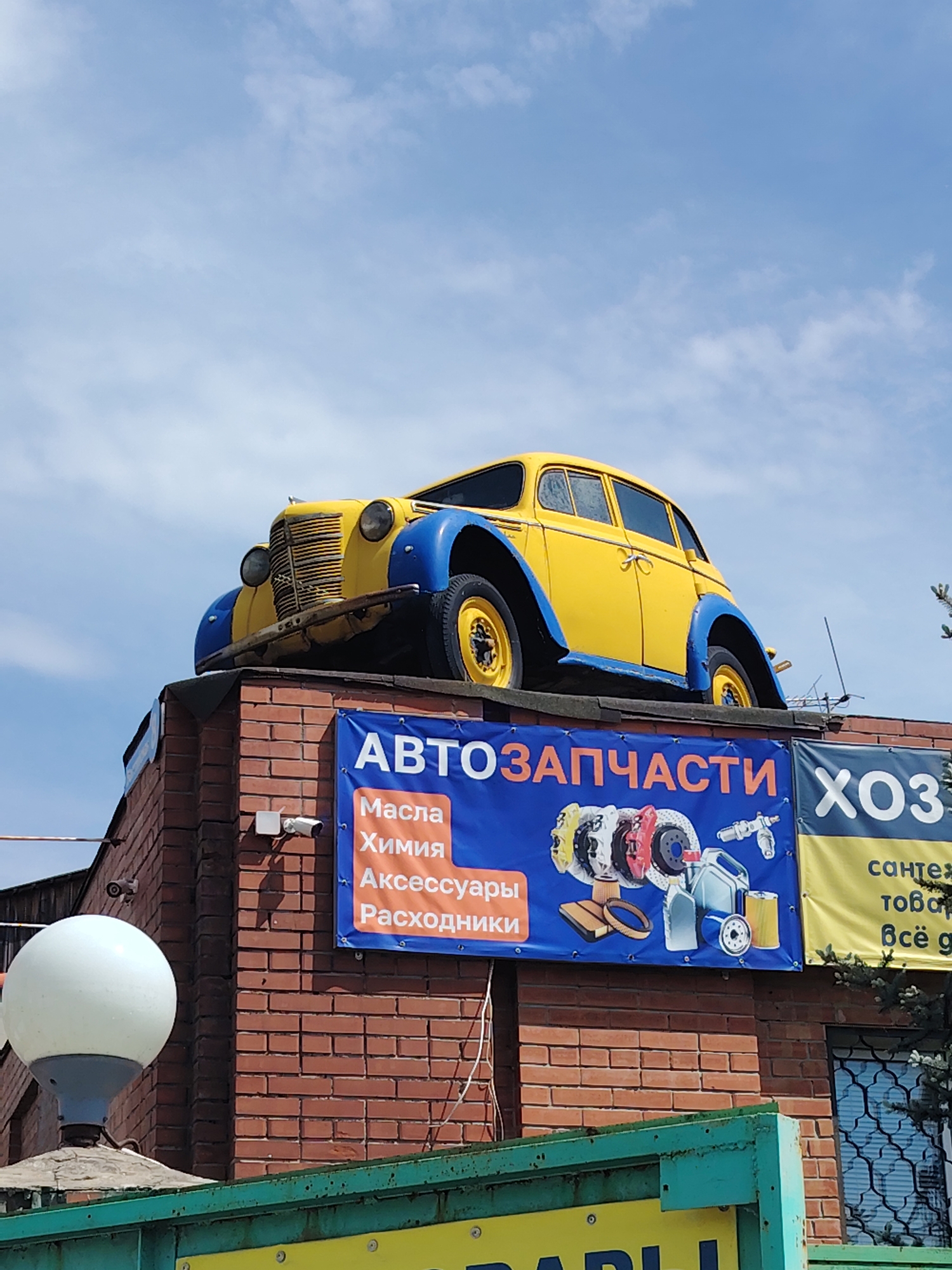 A vintage yellow and blue car is mounted on the roof of a red brick building, with the sky as the background. A blue banner with white lettering reads "АВТОЗАПЧАСТИ" (autoparts) and features illustrations of car parts such as brake discs, filters, and a canister. To the right of this banner, a yellow banner displays "ХОЗ" (household goods) in large white letters and "сантех" (plumbing) and "всё д" (everything for) in smaller text. A white outdoor lamp is positioned to the left of the building, and surveillance cameras are mounted on the brick wall.