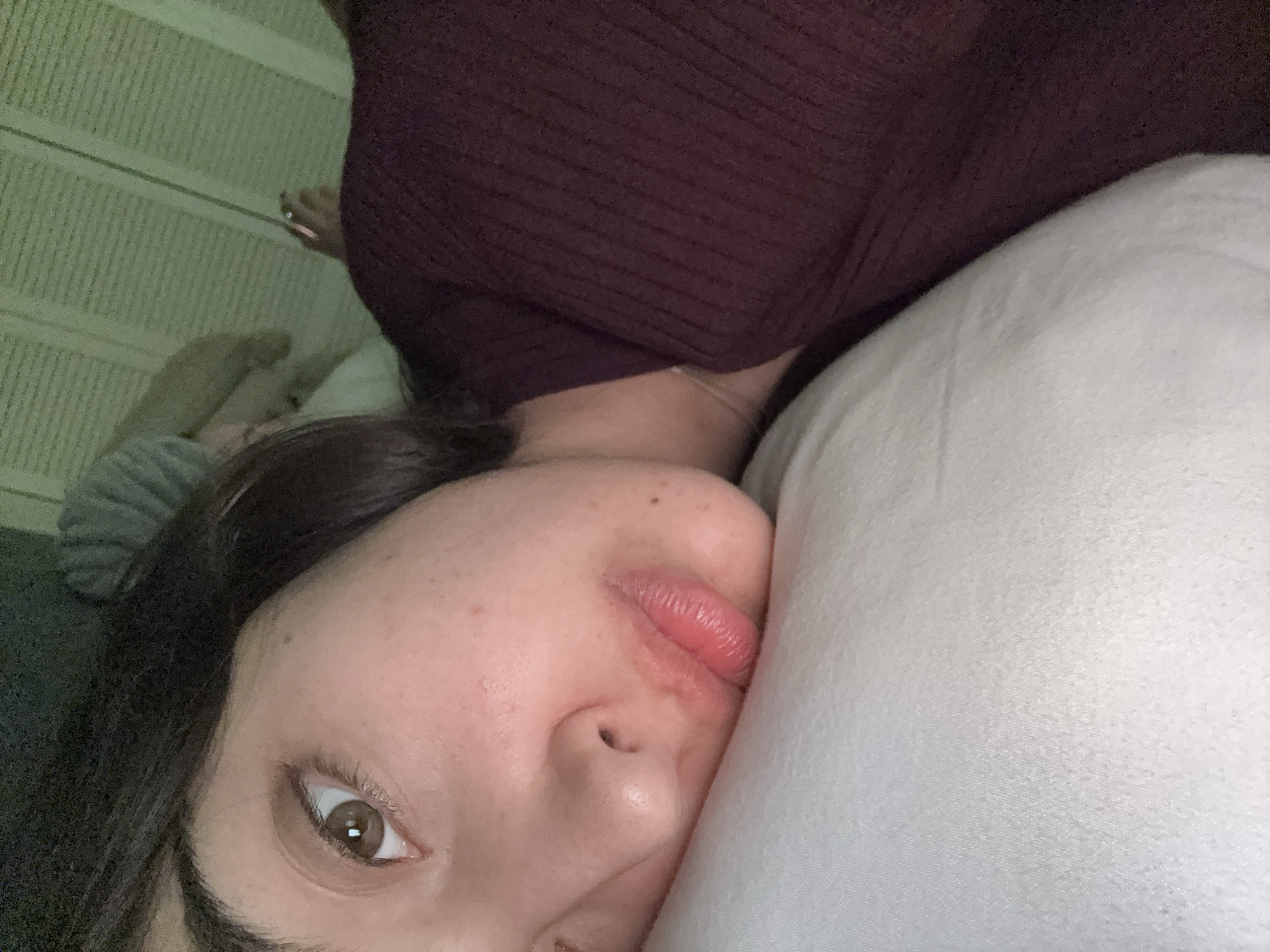A close-up, low-angle, selfie-style shot shows a woman lying on a white pillow, her face pressed against it. She has brown hair and one visible brown eye. Her face has some visible freckles. She is wearing a maroon colored ribbed top. In the background, partial view of what appears to be a wall with vertical lines is visible. The location is not discernible from the image.