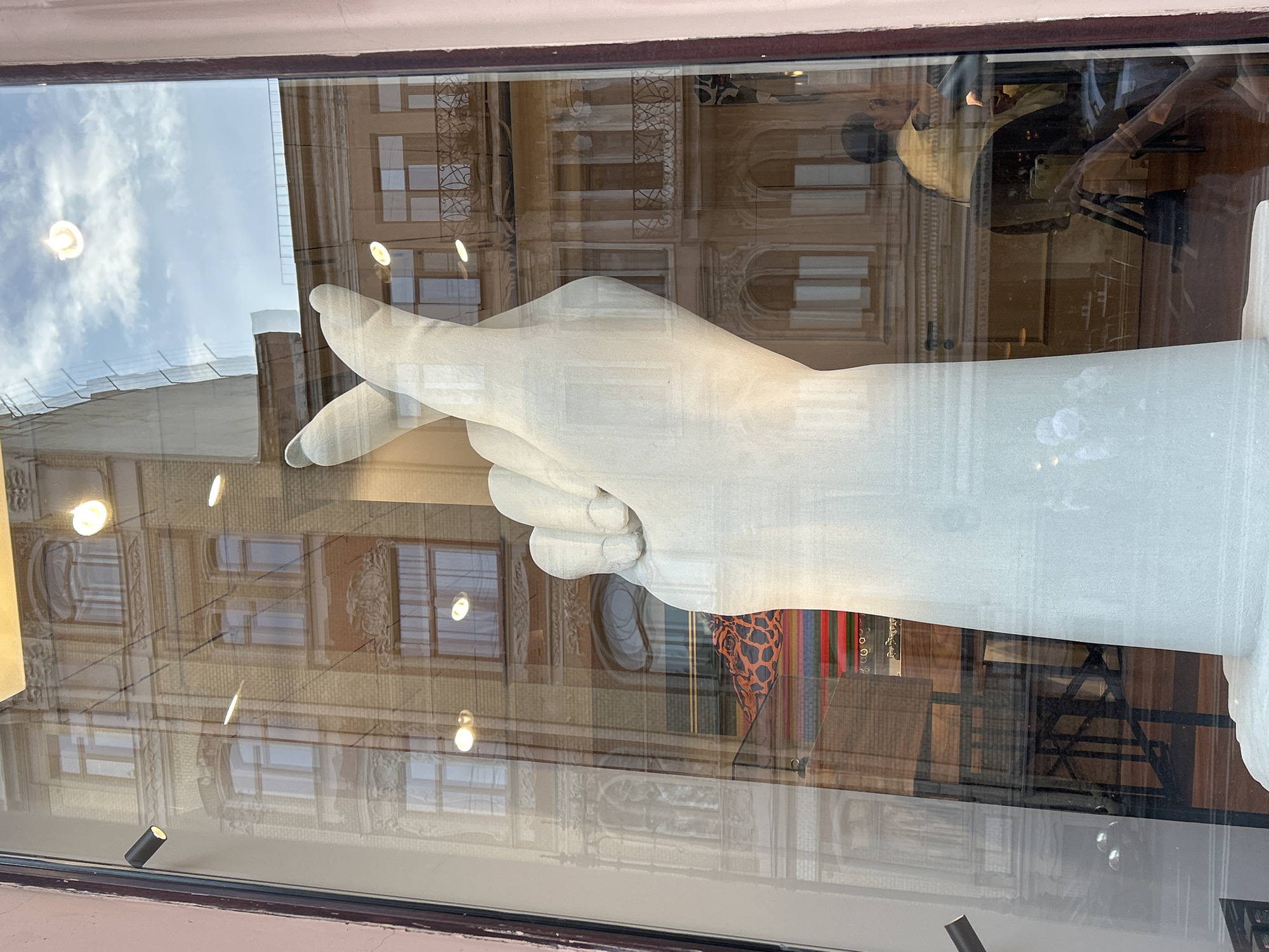 A large, white, stylized sculpture of a human hand, rendered in a textured, matte material, is positioned vertically behind a glass window. The hand forms a "finger heart" gesture, with the thumb and index finger crossed.
Numerous reflections are superimposed on the glass:
* A multi-story building across the street is prominently reflected, featuring a light-colored (possibly yellow or beige) facade with multiple rectangular and arched windows, some with decorative moldings and iron balconies. Architectural details such as cornices and ornate window frames are visible.
* The sky with scattered white clouds is reflected in the upper portion of the glass.
* Several bright, circular light reflections are dispersed across the glass, along with a smaller, dark, cylindrical light fixture reflected on the far left edge.
* On the upper left, a reflection of scaffolding covered with white netting or tarpaulin is visible.
* In the lower right, the reflection of a person wearing a yellow top, leaning on an unseen object, and a hand (likely belonging to the photographer or another person) holding a light-colored mobile phone, are discernible.
Elements visible through the glass, behind the sculpture, include:
* A patterned object or fabric with an orange and black giraffe-like print, partially visible behind the fingers of the hand.
* Below this, horizontal stripes in red, green, and blue are visible, suggesting another display element or fabric.
* Faint outlines of dark furniture (tables and chairs) and a wooden floor can be seen further in the background, indicating an interior space.
The scene is framed by a reddish-pink wall on the left and a darker, possibly wooden, frame on the right.
Location in the city of Unknown, Unknown.