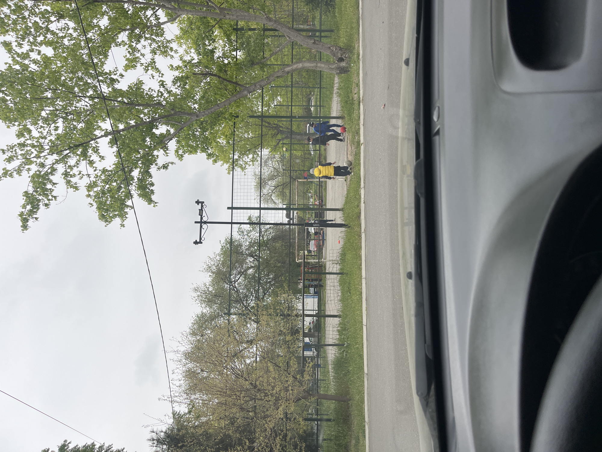 The image, taken from inside a vehicle, shows a scene in Kaspiysk, Russia. A chain-link fence runs parallel to the road on the right. Two trees frame the left side of the fence, with visible foliage. Three people are on the other side of the fence. One individual wears a yellow shirt, and two others are dressed in blue. All three appear to be interacting with a soccer ball on a grassy area. Overhead power lines are also visible. The weather appears overcast. The vehicle's interior is visible in the foreground.