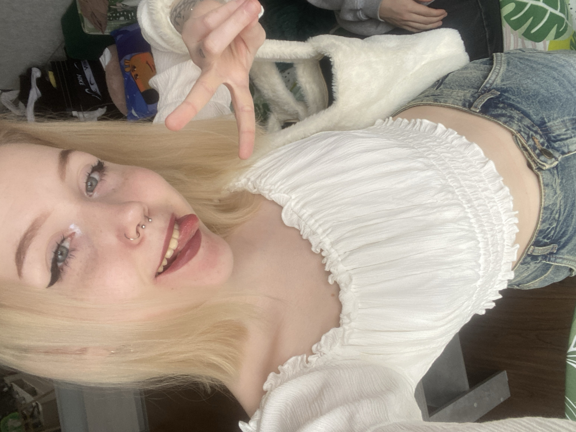 A young woman with light blonde hair is positioned in a selfie, smiling with her tongue extended and making a peace sign with her fingers. She has light skin, visible freckles, winged eyeliner, and a pierced nose. She is wearing a white, off-the-shoulder shirt with a shirred neckline and denim shorts. A fluffy white object, possibly a purse, is in the background. Other visible objects include clothing and a leaf-patterned item. No information is available that relates this image to Izhevsk, Russia.