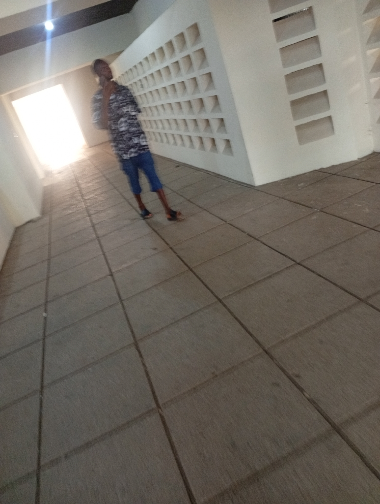 A young Black male wearing a black and white patterned short-sleeved shirt, blue shorts, and sandals stands in a tiled hallway with a bright light source at the far end. The hallway has a textured white wall with a grid of rectangular openings on the right side. The floor is tiled with large, square gray tiles. The lighting suggests it is daytime. The location is identified as the University of Jos, Nigeria.