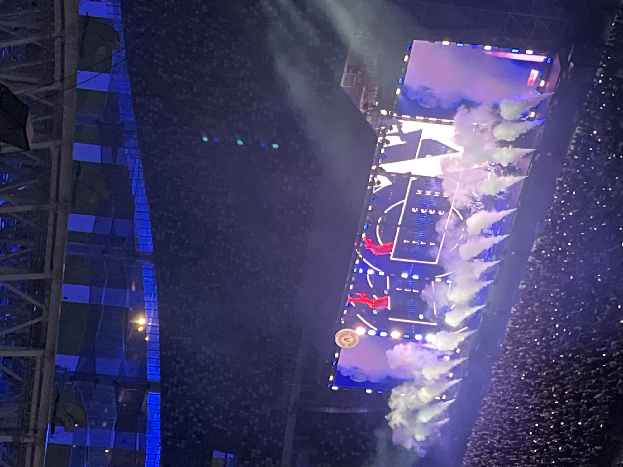 The image depicts a large-scale concert event held within a stadium. The focal point is a massive stage setup, dominated by a large LED screen displaying abstract patterns and graphics. Several red figures are visible on the stage, seemingly performers. Smoke machines are actively deployed, releasing plumes of white and purple smoke across the stage and into the audience area. The stadium is filled with a dense crowd of spectators, many of whom are holding up illuminated mobile phones. The seating area and stadium infrastructure are dimly lit, with blue lighting accentuating the upper structures. The overall atmosphere is one of a lively and energetic live performance. The image is identified as being taken in Nizhny Novgorod, Russia.