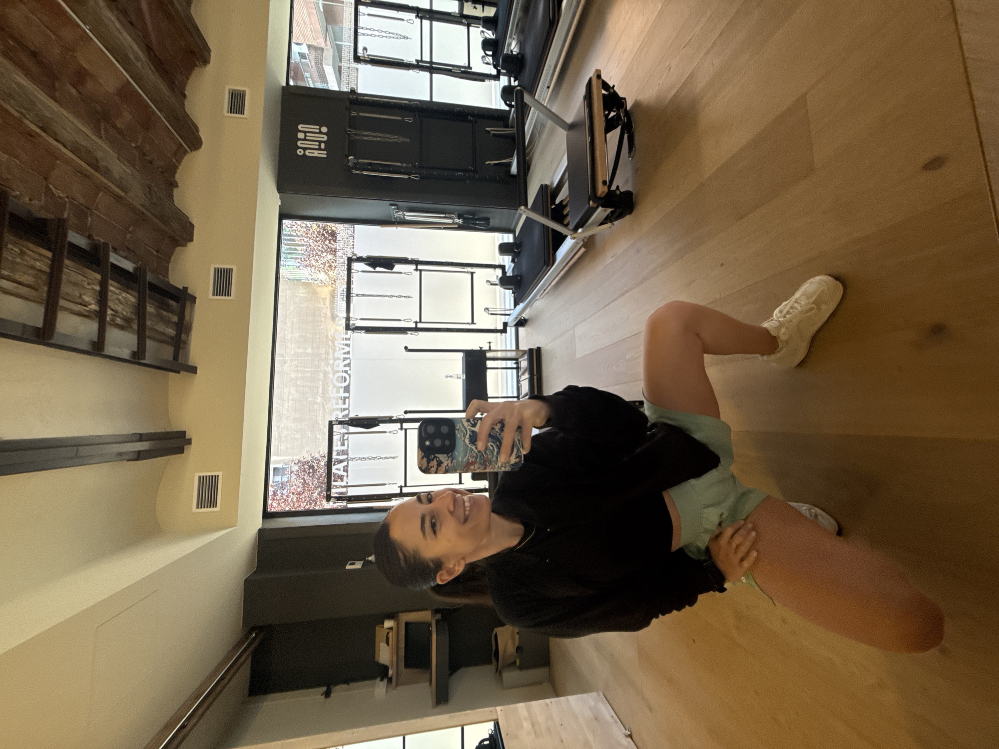 A woman is taking a selfie in a Pilates studio. She is wearing a black zip-up jacket, light green shorts, and white sneakers. She is kneeling on the wooden floor with her left knee bent and her right leg extended. In her right hand, she holds a smartphone with a patterned case. Behind her, several Pilates reformers are visible, and a large window looks out onto a city street. The studio has exposed wooden beams on the ceiling. The studio is in Barcelona, Spain.