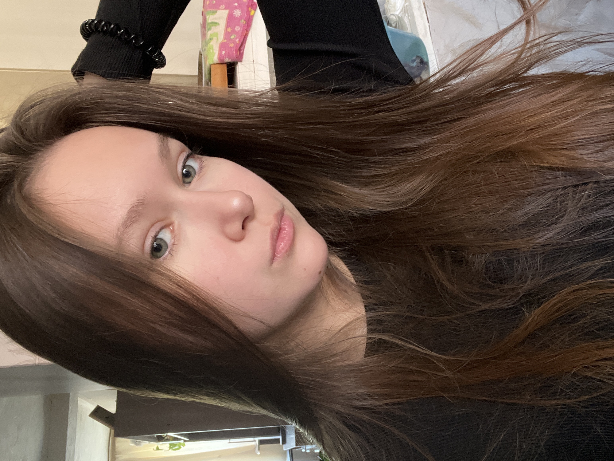 A selfie of a fair-skinned young woman with light brown hair and green eyes, wearing a black long-sleeved shirt and a black beaded bracelet. The background is blurred and includes what appears to be a table with various items on it. The location and city of Chaikovsky, Russia cannot be confirmed from the image alone.