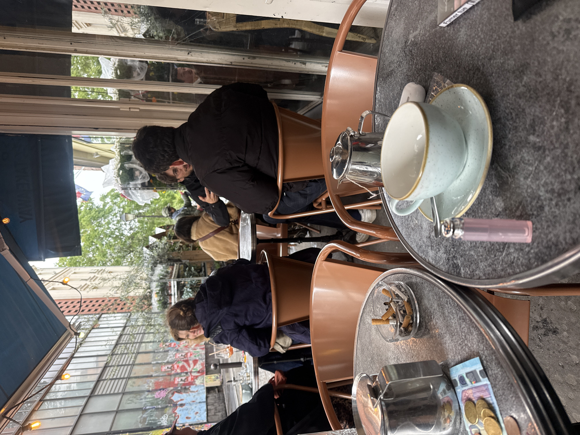 The image shows an outdoor cafe scene, likely in Paris, France, due to the architectural style and ambiance. Several people are seated at small, round tables with metal frames and dark gray surfaces. The tables are arranged close together. Patrons are dressed in casual outerwear, suggesting cool weather.
On the table closest to the viewer, there's a cup and saucer, a metal creamer or sugar container, a lighter, and a small sign or card. The adjacent table has an ashtray filled with cigarette butts, a metallic ashtray, and several coins. The tables are set for customers.
The patrons at the tables appear engaged in conversation or are looking at their phones. The background shows a storefront with large windows reflecting the street. The setting suggests an urban environment, possibly a narrow street or alleyway. Overall, the atmosphere is that of a typical Parisian cafe, with people enjoying drinks and conversation outdoors.