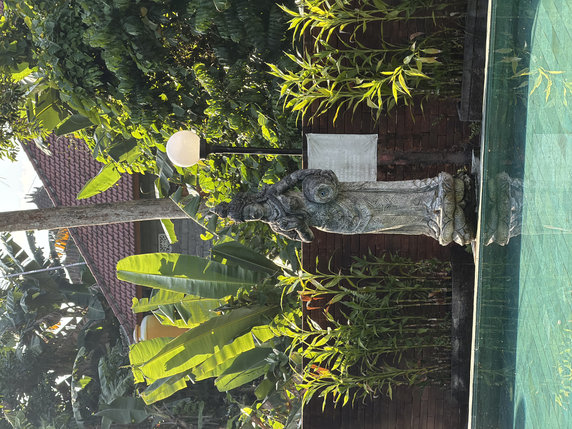A statue of a female deity stands in a garden with lush green vegetation and a swimming pool. The statue is made of stone and is intricately carved, depicting the deity with a crown and holding a pot in one hand. The statue is placed near a brick wall and a streetlight with a white spherical lamp. In the background, banana trees and other tropical plants are visible, along with a part of a building with a tiled roof. The swimming pool in the foreground has clear turquoise water, reflecting the surrounding environment. The scene is bathed in sunlight, creating a serene and tropical ambiance. The location is the district of Ubud in Bali, Indonesia, a region known for its spiritual and natural beauty.