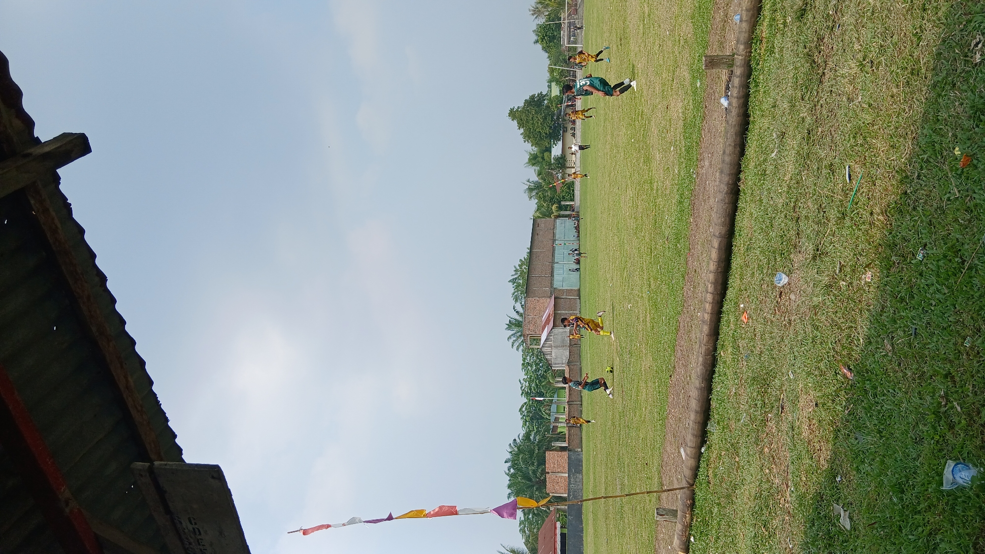 The image shows a soccer game on a grassy field under a bright, cloudy sky. Several young men in athletic uniforms are playing soccer. Some are wearing green and yellow jerseys, while others wear yellow and black. A soccer ball is visible on the field. In the background, there are buildings, palm trees, and fences. A wooden pole with colorful flags is positioned in the foreground on the left side of the image. The field appears to be in a rural or suburban area. Trash is scattered on the grass in the foreground and along the edge of the field. The scene is likely in a village or town in Indonesia, given the presence of palm trees and the style of the buildings.