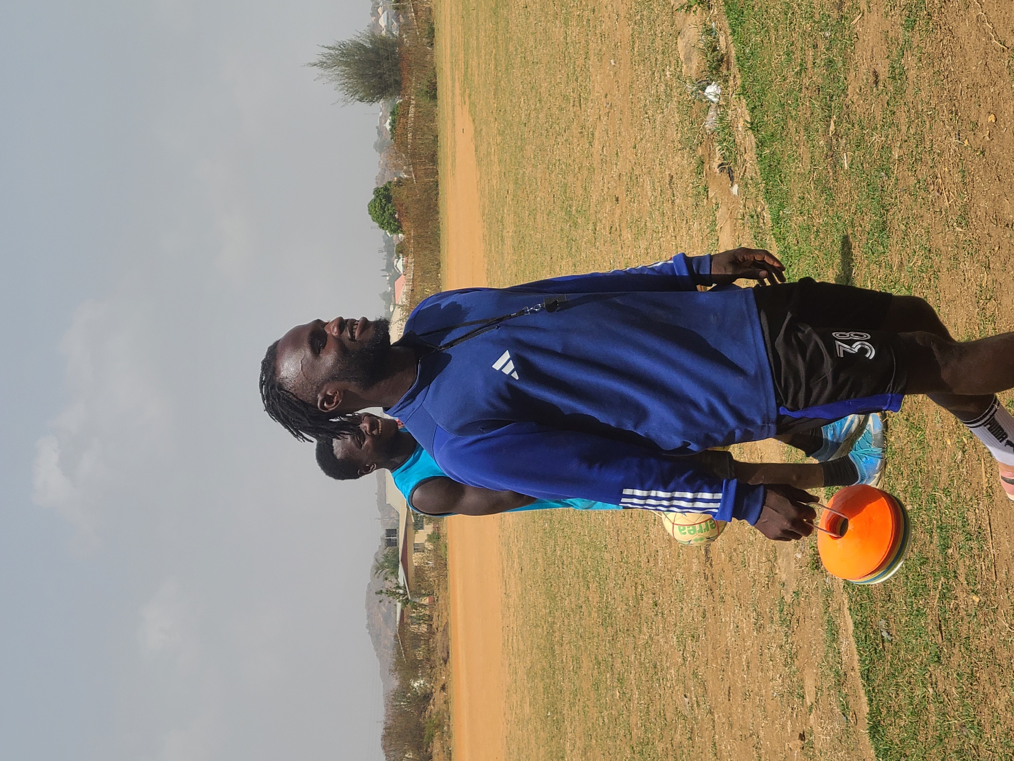 Two Black African men stand on a dirt and grass field in Barikin Jirga Summa, Nigeria. One man, wearing a blue long-sleeved shirt and black shorts with the number "38" on them, holds a stack of orange cones. A soccer ball is near his feet. The second man, slightly behind him, wears a light blue shirt. The sky is overcast. Buildings and trees are visible in the background.