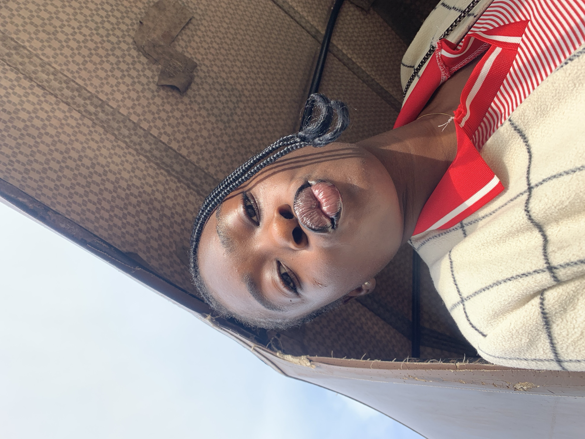 A low-angle, close-up shot captures a young Black woman's face as she poses for a selfie. She is positioned inside a vehicle, likely a keke napep, with a patterned fabric ceiling and brown interior visible. The woman has dark skin, black braided hair that trails over her shoulder, and is wearing a red and white striped shirt under a cream-colored plaid jacket. Her lips are pursed, and her eyes are looking towards the camera. The background is a bright sky. The image is illuminated by natural light, casting shadows. The location is within Gura Top, Nigeria.