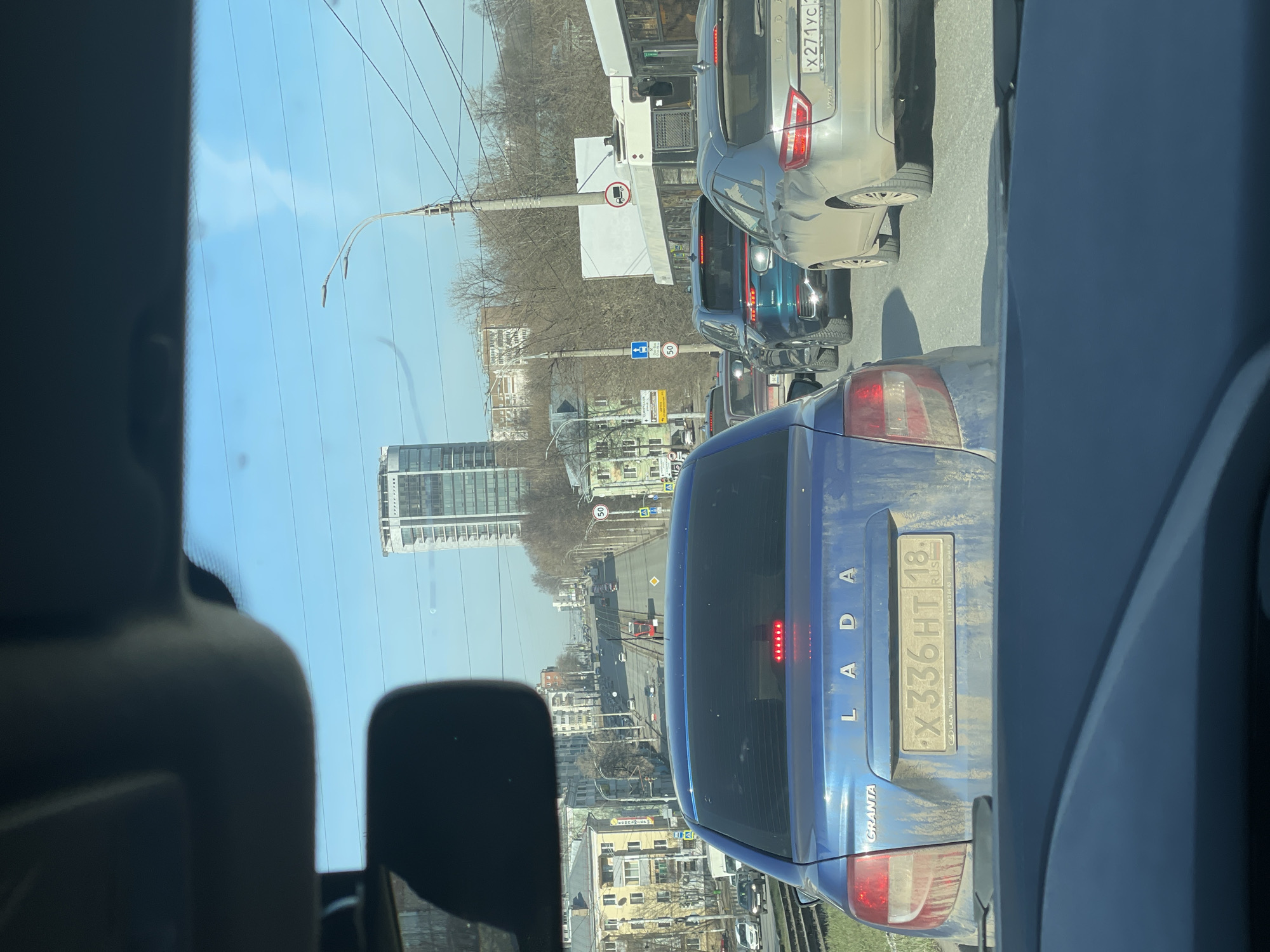 The view from inside a car in Izhevsk, Russia shows traffic on a city street. Buildings of varying heights are on both sides of the road, including a modern high-rise. Streetlights and power lines run overhead. Traffic signs are visible, and a speed limit sign displays "50." In front, a dusty blue Lada Granta bears the license plate X336HT18. Other cars are visible in the traffic ahead.