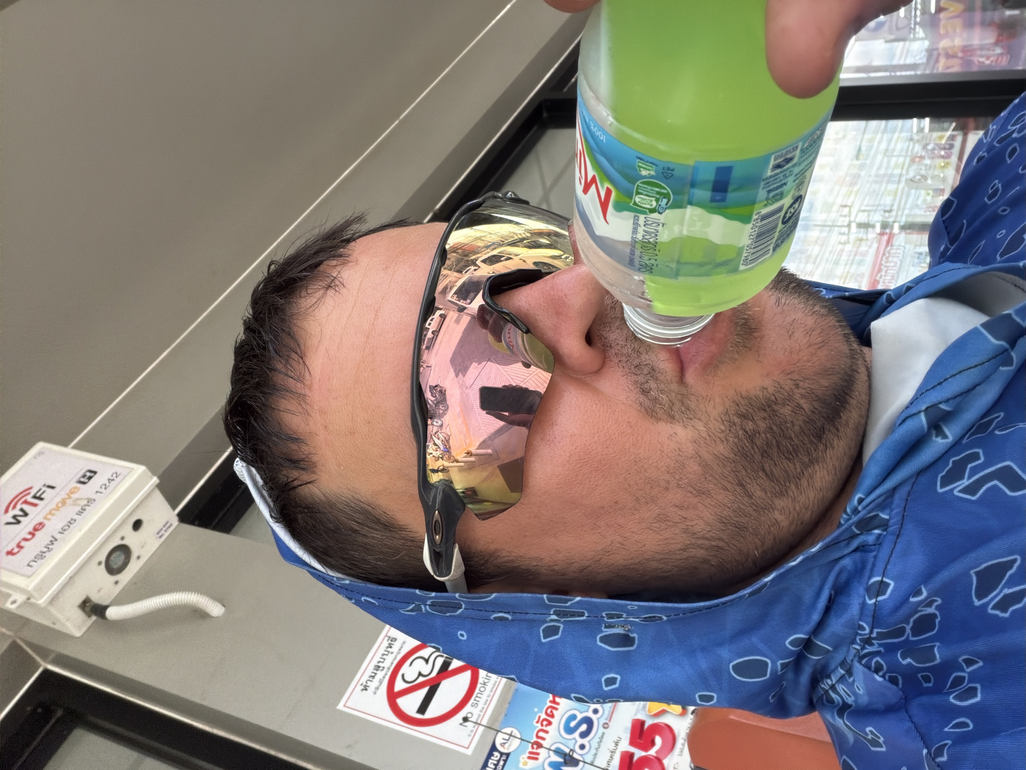 A man wearing a blue hooded sweatshirt and Oakley sunglasses drinks from a green bottle of M-150 energy drink. He is outdoors in Baan Mae Nam, Thailand, with a Wi-Fi hotspot device mounted on a wall above him. A "No Smoking" sign is visible to his left. Reflections in his sunglasses show buildings, vehicles, and pedestrians. The background includes store fronts and power lines.