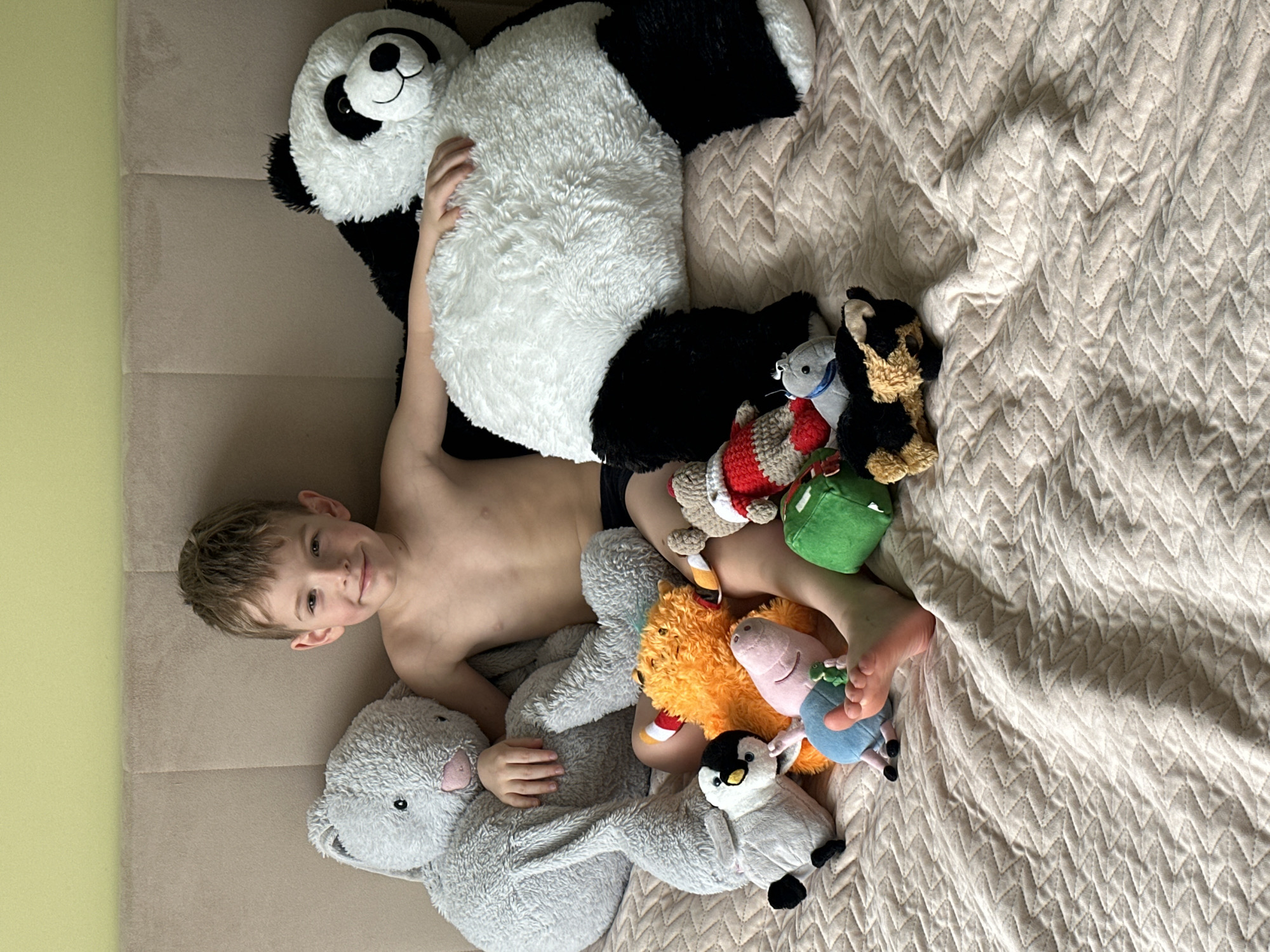 A young boy lies on his back amidst a collection of plush toys, creating a cozy and playful scene indoors, likely in a bedroom or living room in Chaikovsky, Russia. The boy, who appears to be around 5-7 years old, has light brown hair and is smiling warmly at the camera. He is shirtless, wearing only dark underwear or shorts, and appears happy and relaxed.
He is surrounded by numerous stuffed animals. A large black and white panda bear partially covers his upper torso and arms. A grey rabbit or bunny plush is tucked under his left arm. Around his feet and legs, an assortment of smaller toys includes a brown and black dog, a grey mouse, a red and white knitted figure, a green block-shaped toy, an orange fuzzy animal, a pink pig (resembling Peppa Pig), a blue character (possibly George Pig), and a black and white penguin.
The boy and toys rest on a light brown upholstered surface, possibly a sofa or bed, which features two vertical panels. To the right, a light beige quilted blanket with a distinct zigzag pattern covers the rest of the surface. A pale green wall is visible in the background on the far left. The natural lighting suggests it is daytime, contributing to the bright and cheerful atmosphere of the image. No text is visible in the image.