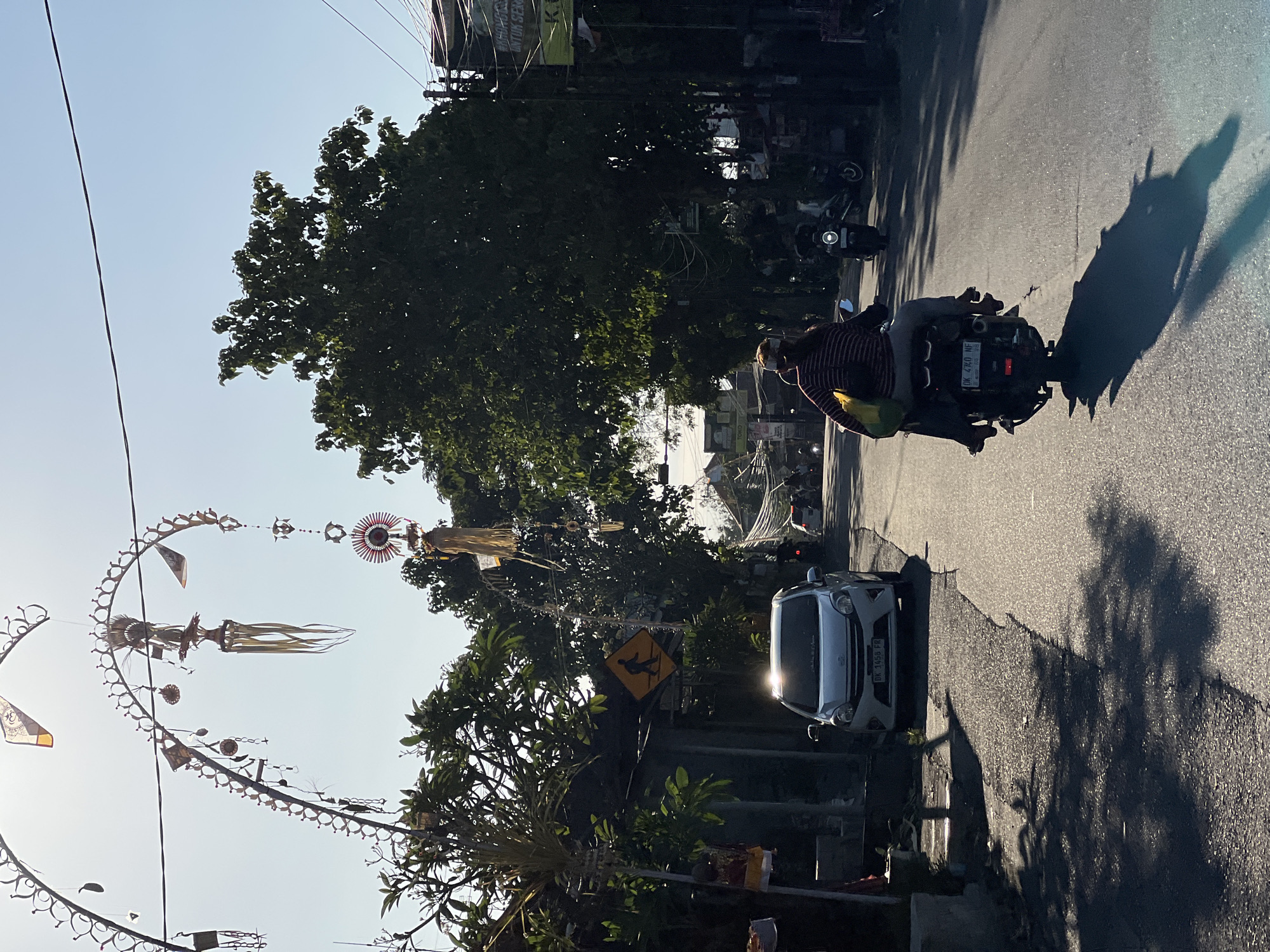 The image depicts a street scene in Kubu, Indonesia, bathed in the warm glow of the late afternoon sun. In the foreground, two individuals are riding on a motorcycle, viewed from behind. The rider wears a striped shirt and a head covering, while the passenger, positioned behind the rider, is seated. The motorcycle itself has a license plate visible, reading "DK 4740 NF".
The road surface is asphalt, showing signs of wear and tear. Shadows of trees and decorative arches are cast across the road, indicating the presence of vegetation and festive decorations. A white car is parked on the left side of the road, with its headlights reflecting the sun. Beside the car, a yellow pedestrian crossing sign is mounted on a pole.
Further down the street, other motorcycles and vehicles are visible, suggesting ongoing traffic. Buildings and trees line both sides of the road. Overhead, decorative arches, adorned with flags and streamers, span across the street, possibly part of a local celebration or cultural display. The sky is clear and blue, contributing to the bright, sunny atmosphere.