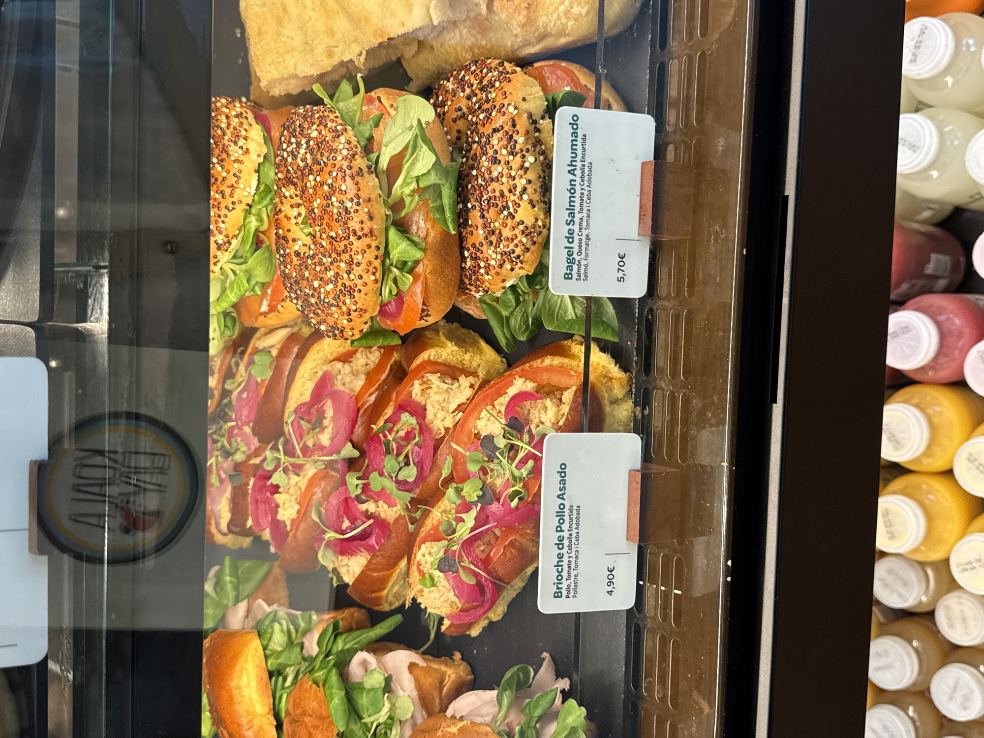 An image captures a food display case, likely in a cafe or deli, presenting various sandwiches and bottled beverages. The image is rotated 90 degrees counter-clockwise, showing the items from a top-down perspective within the display.
The central focus is on two rows of sandwiches. The top row features several bagels, characterized by an abundance of seeds on their crusts. They are filled with salmon, fresh green lettuce or sprouts, and possibly other ingredients, appearing as "Bagel de Salmón Ahumado." Below these, a row of open-faced brioche sandwiches or similar bread items is displayed. These are topped with shredded chicken, slices of red tomato, vibrant pink pickled onions, and green microgreens, identified as "Brioche de Pollo Asado." Further food items, possibly bread or focaccia, are visible at the top and bottom edges of the display.
To the right, a stack of bottled beverages, likely juices or smoothies, are visible, featuring yellow, pink, and red liquids. The display case has a dark interior and a reflective glass front.
Visible text includes two price tags. One for the bagels reads: "Bagel de Salmón Ahumado," with ingredients "Salmón, Queso Crema, Tomate y Cebolla Encurtida" (Spanish) and "Salmó, Formatge, Tomaca i Ceba Adobada" (Catalan), priced at "5,70€." The second tag for the brioche reads: "Brioche de Pollo Asado," with ingredients "Pollo, Tomate y Cebolla Encurtida" (Spanish) and "Pollastre, Tomaca i Ceba Adobada" (Catalan), priced at "4,90€." A circular reflection with partial text like "A JAI" is faintly visible on the left.