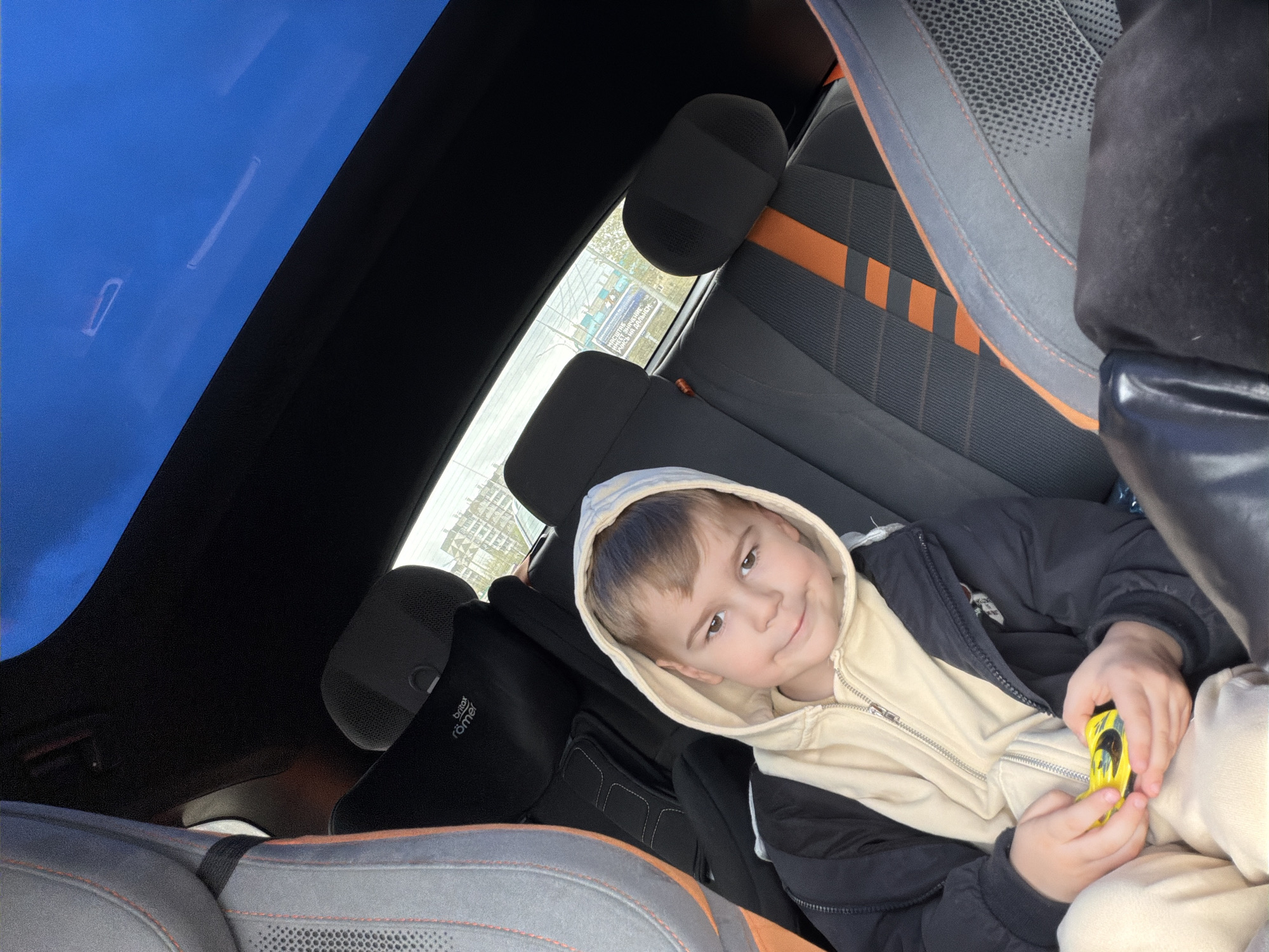 A young boy sits in the back seat of a car. The car has a sunroof with a view of a bright blue sky. The car seats are dark gray with orange accents. The boy wears a beige hooded sweatshirt under a dark jacket and holds a yellow toy car. Outside the car window, a sign in Russian can be seen, placing the location in Krasnoyarsk, Russia.