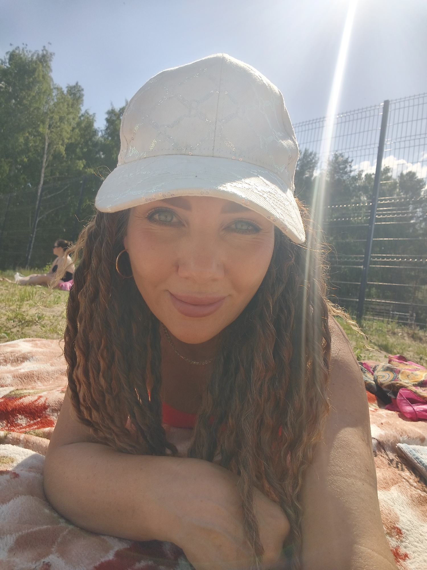 A woman lies on a blanket outdoors. She wears a light-colored cap and has curly brown hair. Her arms rest on the blanket, and she looks toward the camera. In the background, grass, trees, and a wire fence are visible. A person lies on the grass in the distance. Sunlight illuminates the scene.