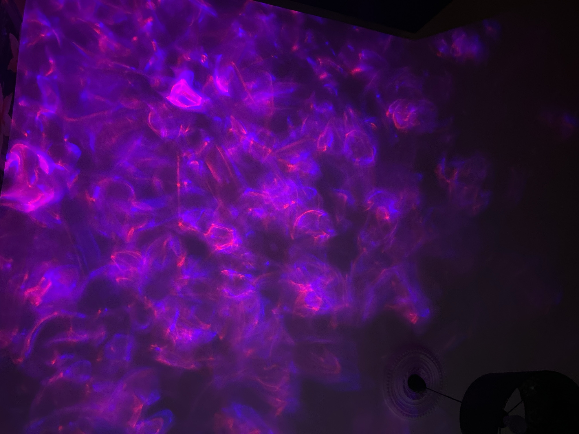 The image displays an indoor scene, likely a ceiling or wall, illuminated by a vibrant and abstract light projection. Dynamic patterns of purple, magenta, and fuchsia light are cast across the surface, creating a flowing, wave-like, or nebulous effect. Brighter hot pink and blue-purple elements are interspersed within the dominant dark purple field, suggesting a moving light source.
The room appears to be dark, indicating that the photograph was taken at night or in a low-light environment to enhance the visibility of the projected lights. In the bottom right corner of the frame, a portion of a ceiling-mounted object is visible. This object is a dark, possibly black, light fixture or fan. Adjacent to it, a decorative, circular ceiling medallion or rosette with intricate concentric patterns is partially discernible.
No people are present in the image. There is no visible text. The scene is an interior in Sofia, Bulgaria, where the artificial lighting creates a distinct ambiance, likely for relaxation or aesthetic appeal.