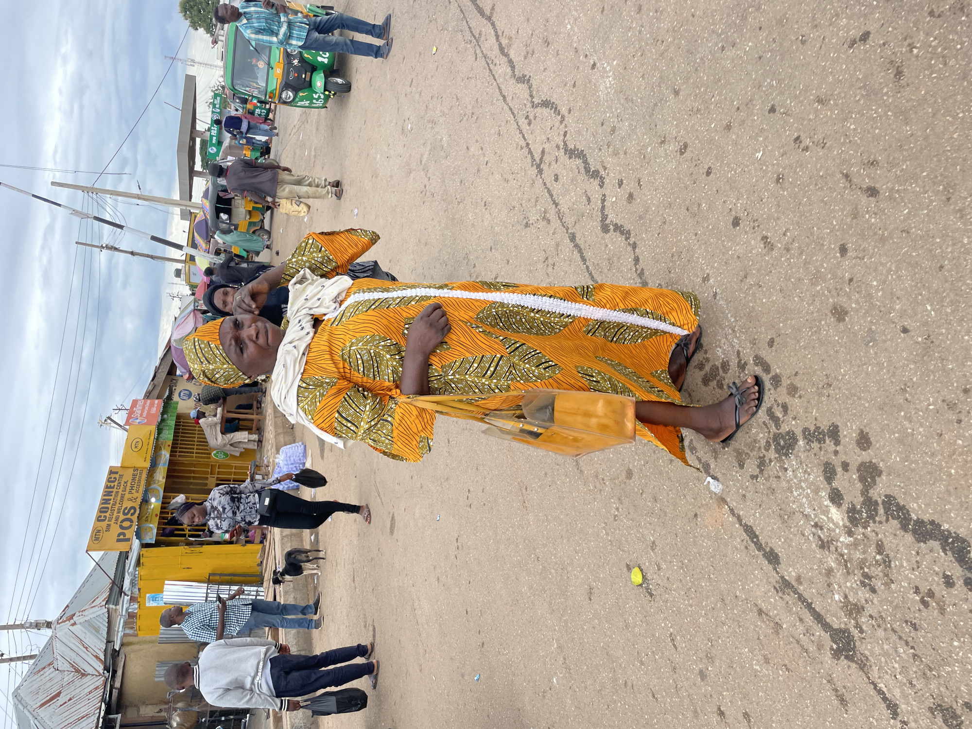 The image depicts a busy street scene in Gura Top, Nigeria. In the foreground, a woman wearing a vibrantly colored orange and green patterned dress and a headscarf is captured mid-stride. She carries a large, transparent yellow tote bag over her right shoulder and has her left hand raised to her face. She is wearing dark sandals. Behind her, the street is populated with various individuals and vehicles. To her left, a young boy in a grey sweatshirt and dark pants walks away from the camera, carrying a black bag. A man in a blue and white checkered shirt stands nearby, looking to his right and holding a mobile phone. A small black dog stands between them. In the background, several yellow and green auto-rickshaws, commonly known as "Keke Napep," are visible, some of which appear to be in motion. A yellow building with signage that reads "MTN CONNECT SIM REGISTRATION CENTRE AND WELCOME BACK POS & PHONES ACCESSORIES" is prominent on the left side of the image. Umbrellas and awnings suggest market stalls. Power lines and poles crisscross the sky, and the overall environment suggests an outdoor, possibly commercial or transportation hub. The ground is paved with asphalt, showing signs of wear and some dark stains. The sky is overcast with a muted light.