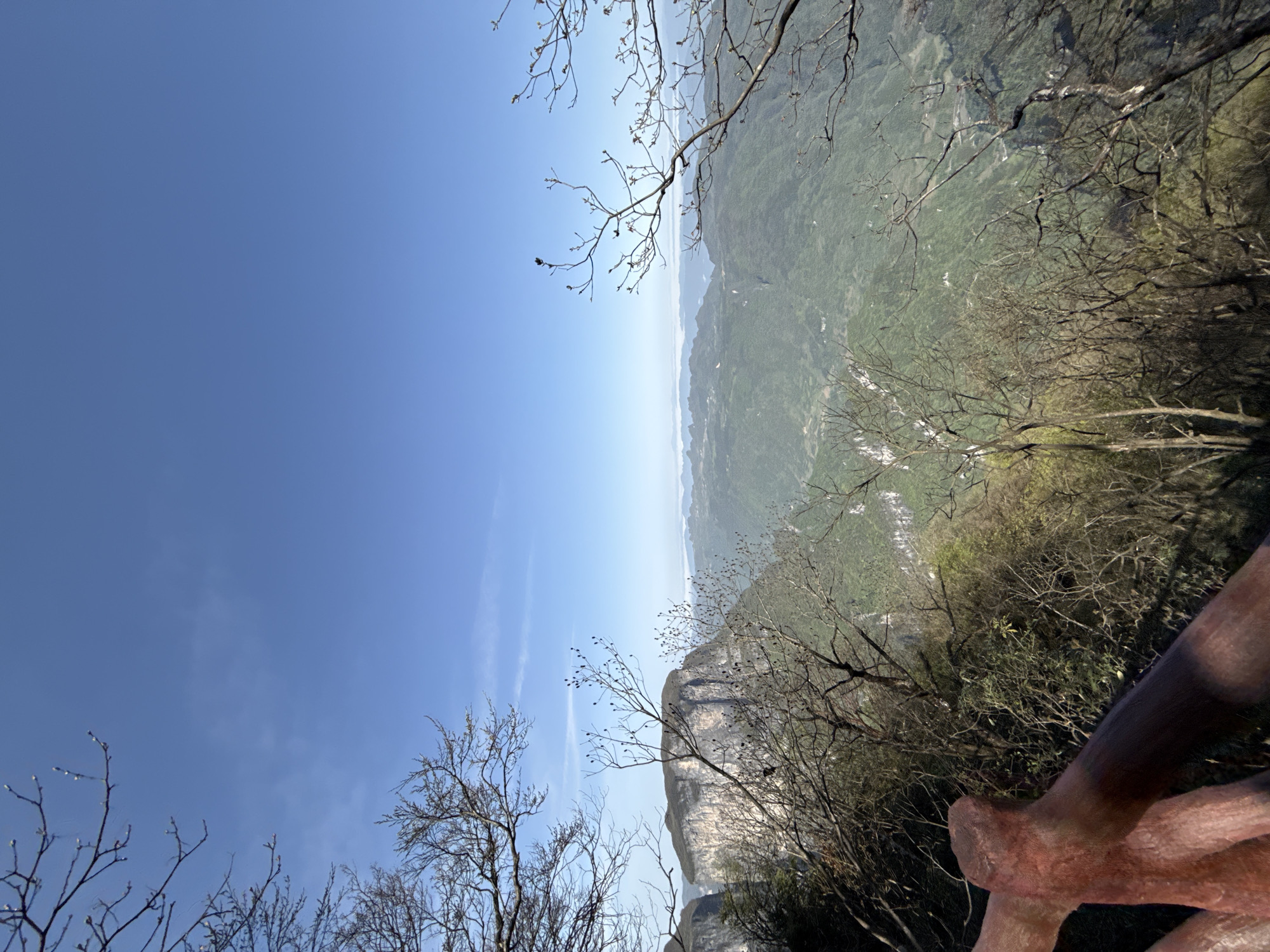 A landscape photograph taken in Yongding District, China, shows a view from an elevated perspective. The sky is a gradient of light blue with wispy clouds. Below, a valley is visible, covered in greenery and interspersed with light-colored areas that might be rivers or settlements. Bare tree branches frame the top and bottom of the scene, and a rocky cliff is visible on the bottom. A reddish-brown wooden structure appears in the lower right corner.
