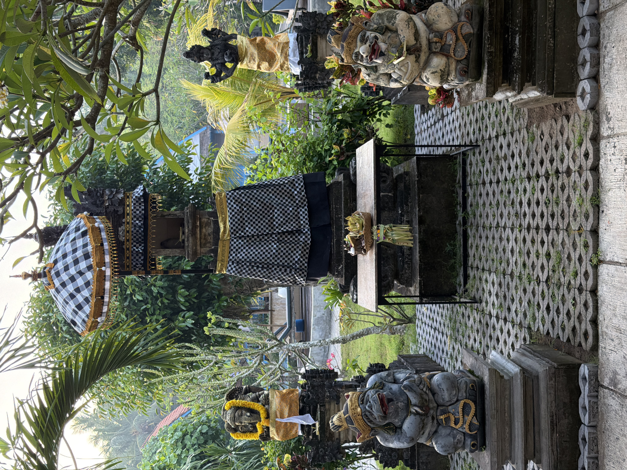 The image captures a courtyard in Toyapakeh, Indonesia. In the center stands a small Balinese shrine, draped in a black and white checkered cloth, with an ornate umbrella shading it. A table holds a small offering of leaves and flowers. Two stone guardian statues, decorated with gold accents, flank the entrance to the shrine. The ground is covered in interlocking concrete pavers. Lush tropical foliage surrounds the shrine, with glimpses of buildings in the background. The scene is captured on a slightly overcast day.