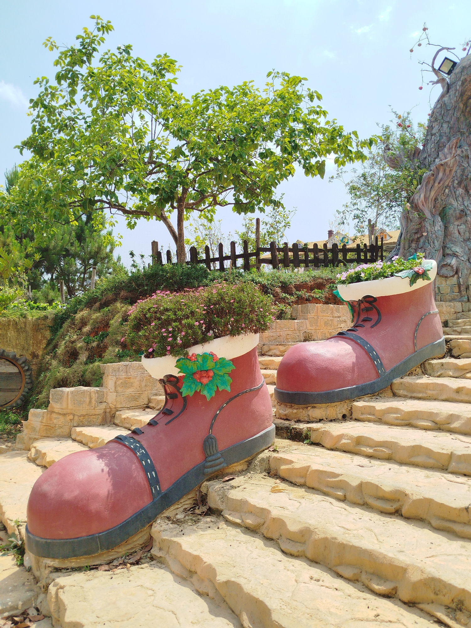 The scene is a whimsical garden display on stone steps, located in what is reported to be Lang Biang - Da Lat, Vietnam. Two oversized, reddish-brown boot sculptures serve as planters. The boot closer to the viewer is decorated with a painted green and red design, possibly representing foliage. The planters are filled with vibrant pink and white flowers, which are overflowing from the boot tops.
A small, rustic wooden fence and lush greenery fill the background. A tree with green foliage stands at the top of the steps, with blue sky visible above it. To the right, there's an artificial tree with decorations, appearing to include apples. The overall impression is of a playful, colorful, and slightly surreal garden setting.