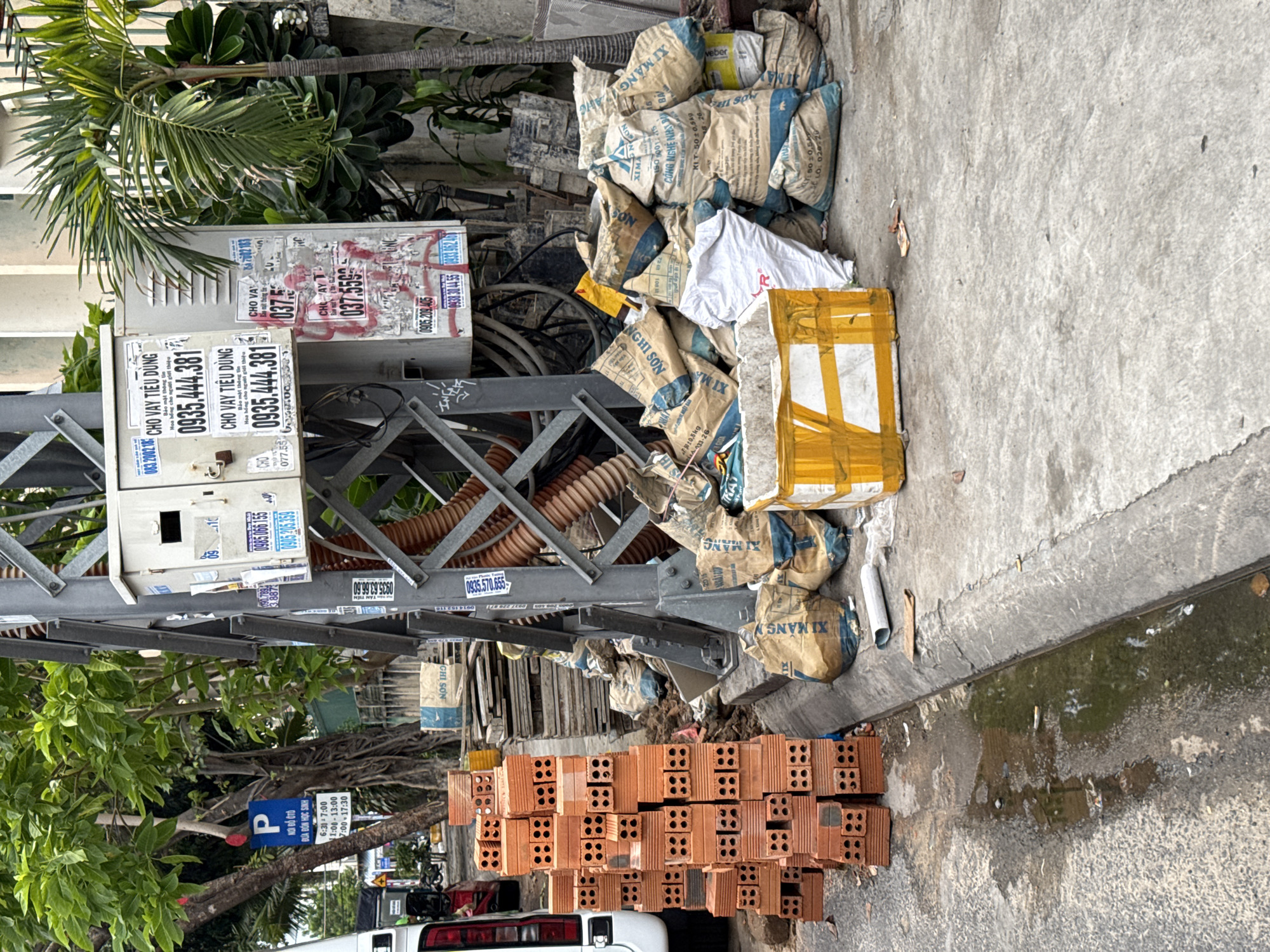 The image depicts a street scene in Nha Trang, Vietnam, with a focus on construction materials and utility infrastructure. In the foreground, a stack of red bricks is piled on the asphalt, adjacent to a concrete curb. Beyond the curb, several large sacks, likely containing cement or other building materials, are piled haphazardly. Some of these sacks have blue and yellow markings with Vietnamese text. A white plastic bag is also visible amongst the sacks. To the left, a portion of a white vehicle is visible.
Dominating the background is a metal utility pole with a complex arrangement of wires and pipes. Attached to the pole is a grey electrical box with several posters and advertisements adhered to it, many of which feature phone numbers and Vietnamese text, suggesting commercial services. One poster, prominently displayed, reads "CHO VAY TIEU DUNG" (Consumer Loan). A large, corrugated orange pipe runs vertically along the utility pole.
The environment appears to be an urban setting with a mix of vegetation, including a lush green tree on the left and a palm tree on the right, suggesting a tropical climate. The ground is a mix of asphalt and concrete, with evidence of recent rain or water on the asphalt, forming a small puddle in the gutter. The overall impression is one of an active construction or maintenance area on a city street.