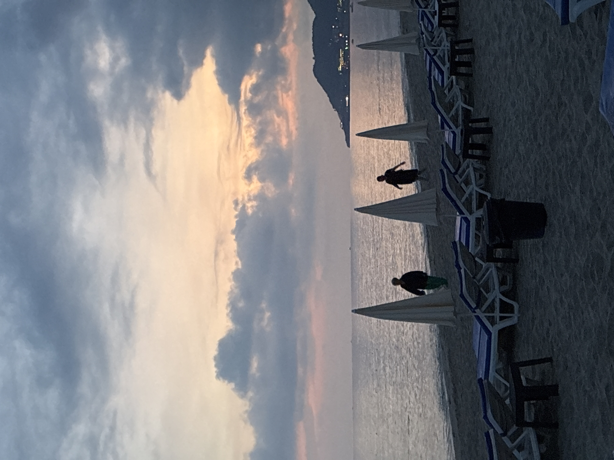 This image depicts a serene beach scene in Alanya, Turkey, at dusk. The sky is a canvas of soft hues, transitioning from a pale peach near the horizon to deeper blues and grays as they merge with dramatic clouds. A gentle sea laps at the shore, reflecting the muted light.
On the sandy beach, several empty lounge chairs, arranged in neat rows, hint at a day's end of relaxation. White beach umbrellas, some partially obscuring the view, stand sentinel along the shoreline. In the distance, a dark, tree-covered hill rises, its silhouette punctuated by distant lights, suggesting the presence of a town or resort.
Two figures are visible walking near the water's edge. One, further back, is a person in dark clothing walking towards the right side of the frame. Closer to the foreground, another individual, also in dark attire with what appears to be green pants, is walking towards the left. They are positioned between two beach umbrellas. A dark trash receptacle is present in the lower right of the frame. The overall atmosphere is tranquil, with the fading light creating a sense of calm and reflection.