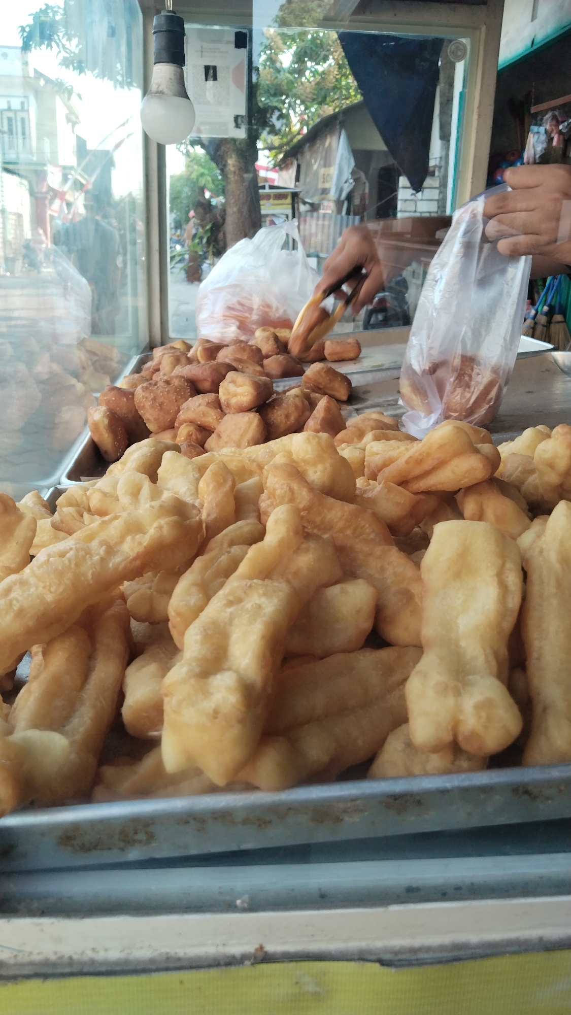 This is a close-up, eye-level shot of a food stall, likely in Peganden, Indonesia, specializing in fried dough pastries. In the foreground, a large quantity of golden-brown, fried dough sticks, some twisted and elongated, are piled high in a metal tray. Behind them, a second tray holds smaller, bite-sized, fried dough pieces, some with a textured surface. To the right, a person's hands are visible, holding tongs and picking up a piece of fried dough from a plastic bag, and another plastic bag filled with pastries sits nearby. In the background, through a glass enclosure, a glimpse of the street and trees is visible, along with a simple stall structure. A bare lightbulb hangs from the ceiling of the stall.