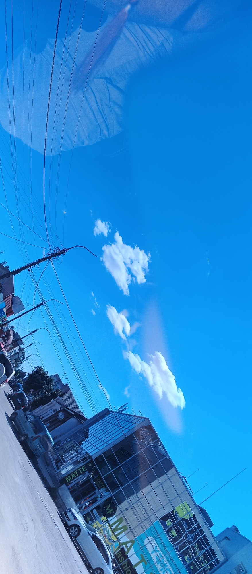 A low-angle shot captures a bright blue sky filled with scattered white clouds. Overhead, a network of power lines crisscrosses the frame, casting faint shadows. In the foreground, a modern building with a glass facade reflects the sky and surrounding environment. The building displays signage, with "MATTE Fashion" prominently visible. A white car is parked on the street in front of the building. The scene appears to be a street in the city of Smila, Ukraine, bathed in sunlight.