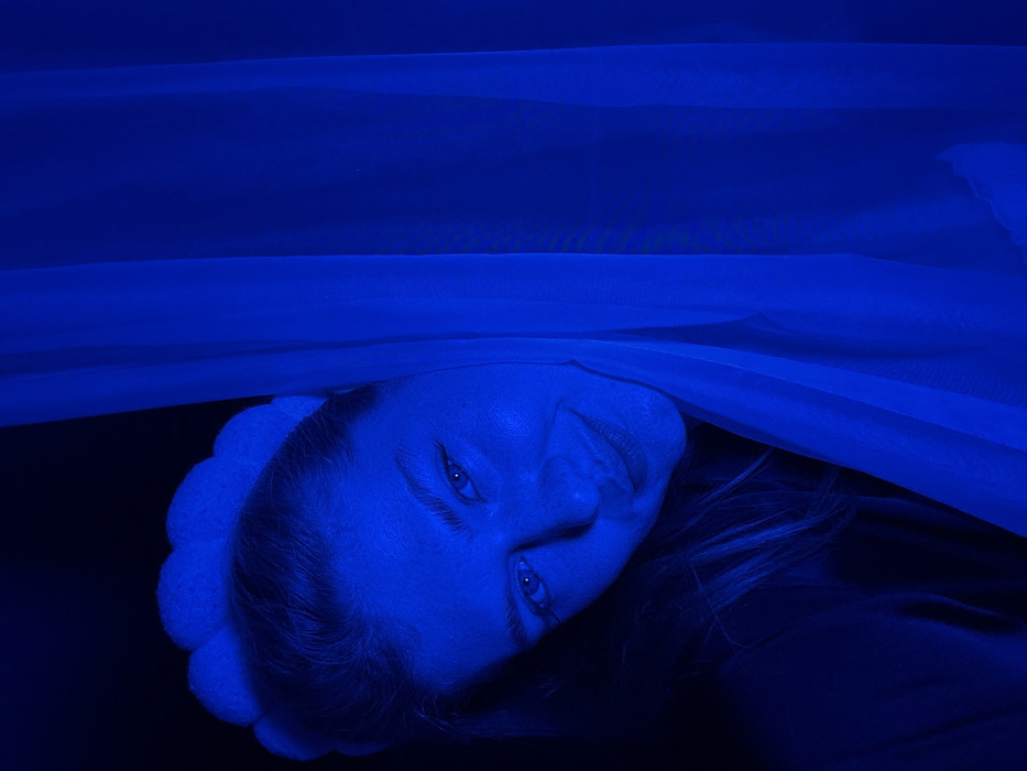 A woman's face, bathed in intense blue light, dominates the image. She is positioned at an angle, tilted towards the left. A white, semi-transparent fabric drapes over her and extends across the top of the frame. Her eyes are open, and she appears to be smiling faintly. The background is dark and indistinct. She has a knitted hairband with bobbles on her head. Parts of her hair are visible.