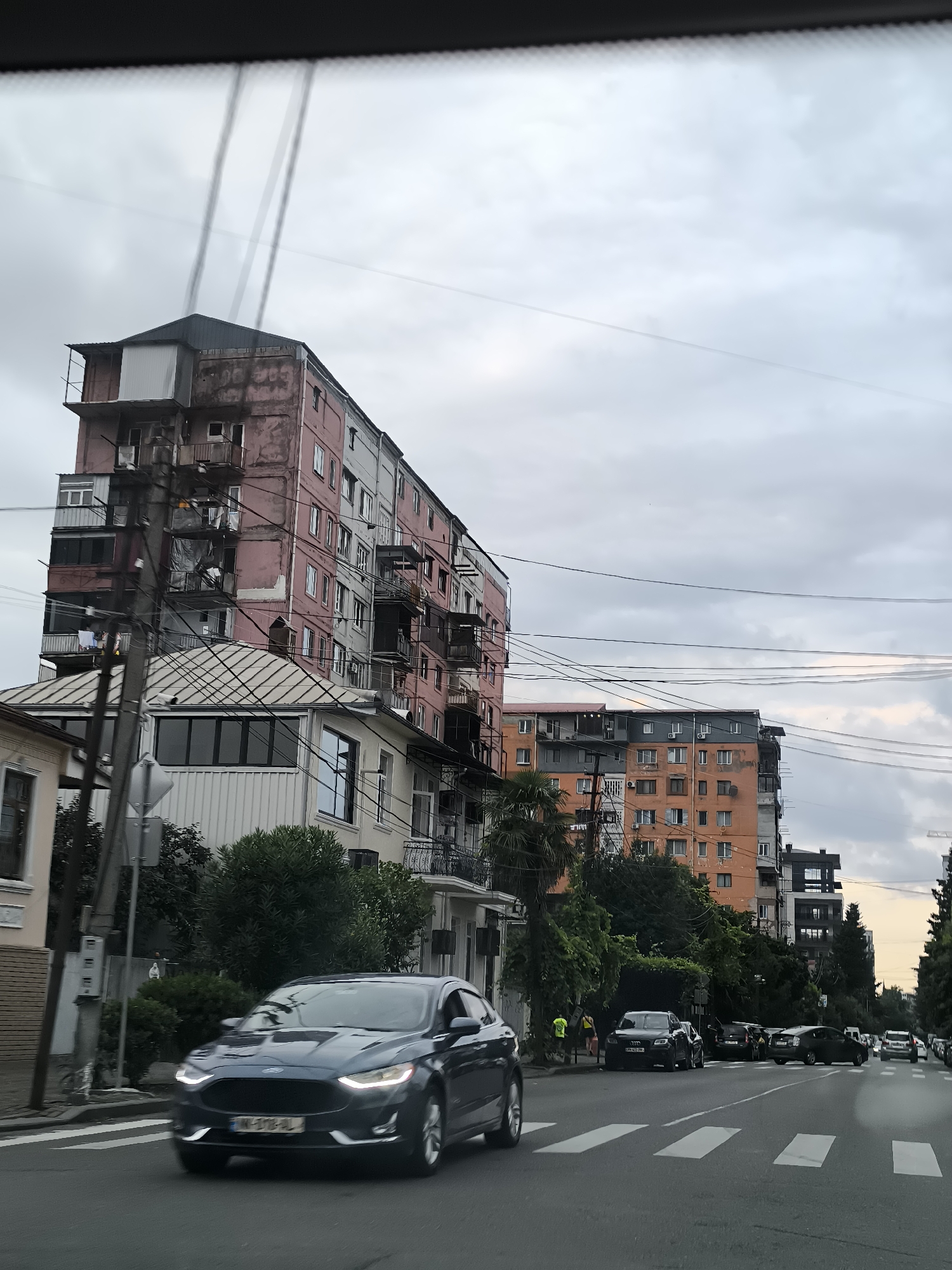 A dark blue Ford sedan drives on a street in Batumi, Georgia, passing by residential buildings. The buildings are multi-story apartment blocks with a mix of colors, some appearing weathered. The architecture is typical of Soviet-era construction, with balconies and visible external wiring. Several other cars are parked along the street and on the road. There are pedestrians on the sidewalk, some wearing bright yellow vests. The sky is overcast with a muted, cloudy appearance. Power lines crisscross the sky and are visible in front of the buildings. The street has white pedestrian crossings. The overall scene depicts an urban environment with everyday street activity.