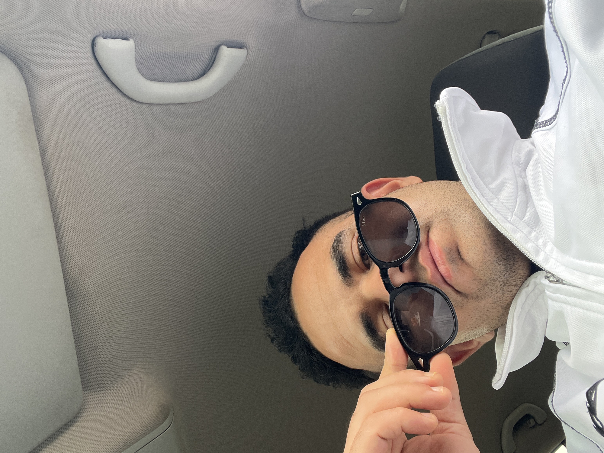 An overhead shot captures a man inside a car, looking upwards and slightly to his left. The man, with dark hair, dark eyebrows, and light stubble, occupies the lower right quadrant of the image. He wears dark sunglasses with prominent black frames, and his right hand gently touches the bottom rim of the right lens. The word "Dior" is faintly visible on the temple arm of the sunglasses. He is dressed in a white zippered jacket or shirt, featuring dark stitching along the collar and zipper line.
The setting is clearly a car interior, characterized by the light-colored, textured fabric of the headliner and a white grab handle (assist handle) mounted on the roof. Part of a car pillar, covered in the same fabric, is visible on the left, and a dark car seat or headrest is partially seen behind the man's head. The lighting suggests it is daytime. The man appears calm, seemingly posing for a selfie or photo. This scene takes place in Martil, Morocco.