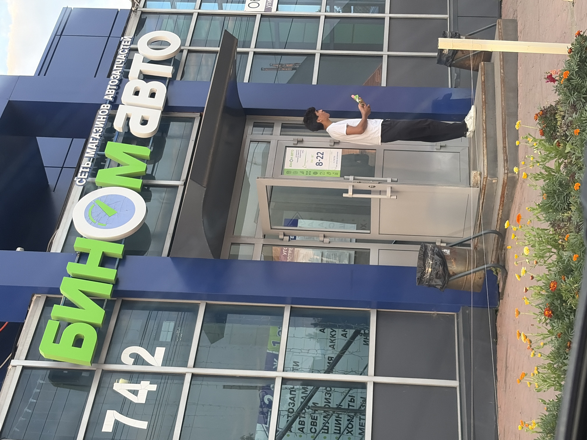 A street-level exterior view captures the entrance of a business named "БИНOМ АВТО" (BINOM AVTO), a chain of auto parts stores, located at house number 742 on a street in Izhevsk, Russia. The store features a bright blue facade with large glass windows. Prominently displayed above the entrance are large, vibrant green and white three-dimensional letters spelling out the business name. Below this, smaller white text reads "СЕТЬ МАГАЗИНОВ АВТОЗАПЧАСТЕЙ" (CHAIN OF AUTO PARTS STORES).
A young man wearing a white t-shirt and black pants stands on the steps leading up to the entrance, holding a small, colorful object in his right hand. He is facing to the right, away from the camera. The entrance itself has a glass door with a silver handle. To the left of the entrance, visible through the glass windows, are reflections and signs indicating various auto parts such as "АВТОЗАПЧАСТИ" (AUTO PARTS), "АВТОЛАМПЫ" (AUTO LAMPS), "СВЕЧИ" (SPARK PLUGS), "ШУМОИЗОЛЯЦИЯ" (SOUNDPROOFING), "ШИНЫ" (TIRES), "ХОМУТЫ" (HOSE CLAMPS), and "МЕТИЗЫ" (FASTENERS).
To the right of the entrance, there is a trash can with a black plastic bag. In the foreground, a planter box filled with orange and yellow flowers adds a touch of color to the scene. The sky above appears to be partly cloudy. The overall impression is a commercial street scene on an overcast day.