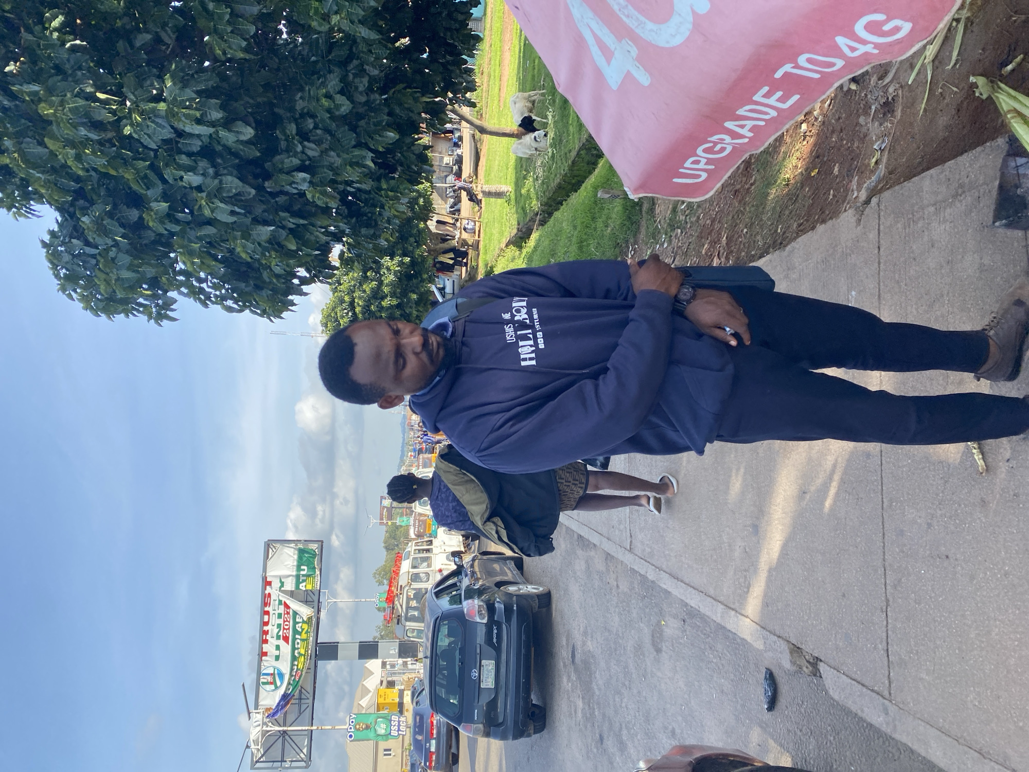 A medium shot captures a man standing on a sidewalk in Gura Top, Nigeria. He is wearing a dark blue hooded sweatshirt and dark pants, with a watch on his left wrist. The man is looking down, with his arms clasped in front of him. To his left, a black Toyota Matrix car is parked on the side of the road. Further down the road, several other vehicles and pedestrians are visible. Behind the man, a large green tree stands, and in the background, a billboard advertising "TRUST FOR UNITY 2021" is visible. Two sheep, one black and white and one white, are grazing on a grassy area to the right of the man. A pink banner with white lettering reading "UPGRADE TO 4G" is also present in the foreground to the right. The scene appears to be a street with moderate traffic and commercial activity.
