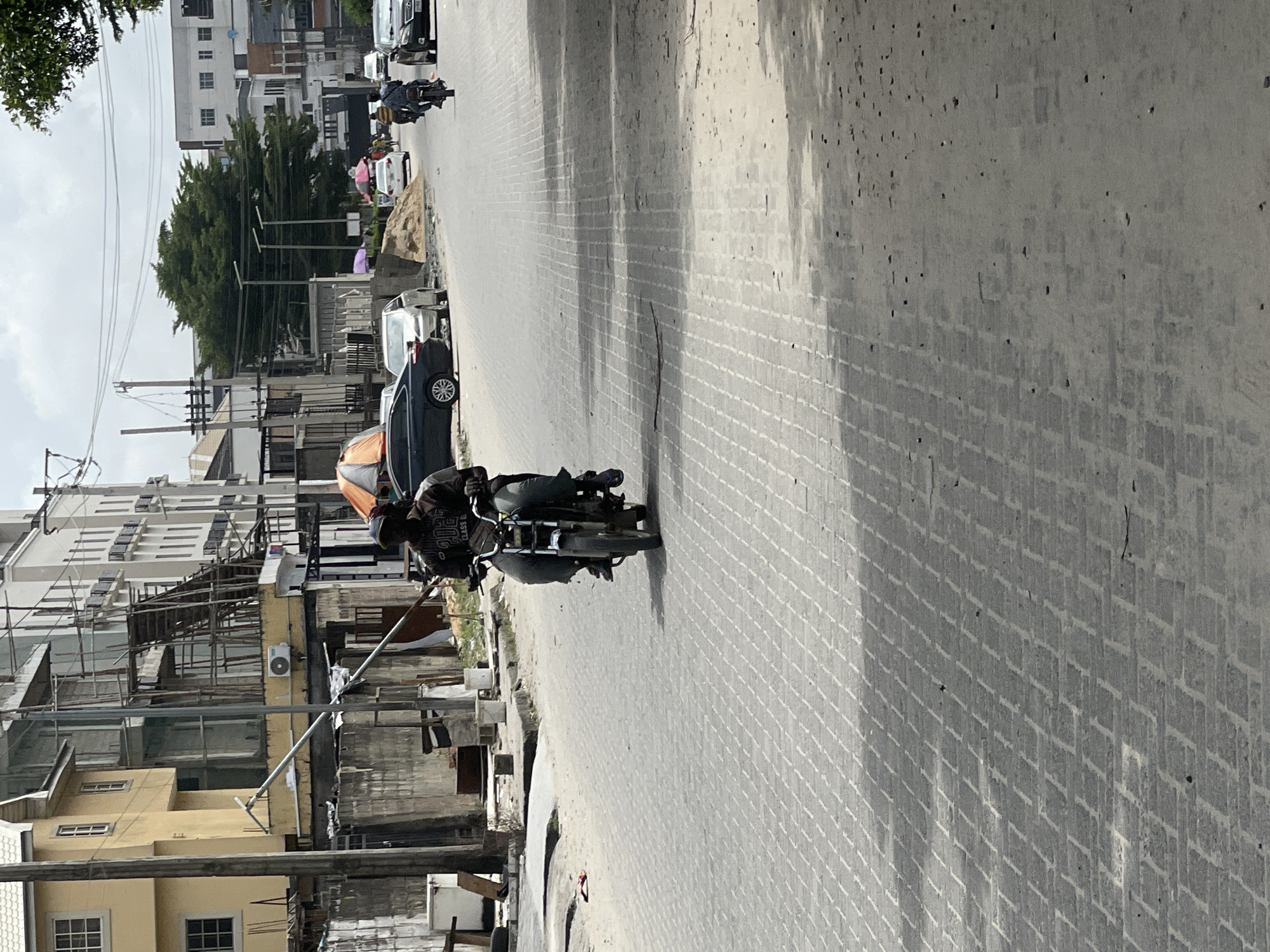 In Igboefon, Nigeria, a motorcyclist is seen riding down a paved street. The street is made of interlocking bricks and is lined with buildings. The buildings appear to be a mix of residential and commercial properties. The sky is overcast. The motorcyclist is wearing a dark shirt and pants, and is carrying a passenger behind him. The motorcycle is a dark color. There are a few cars parked on the side of the road. The scene is depicted in daylight.