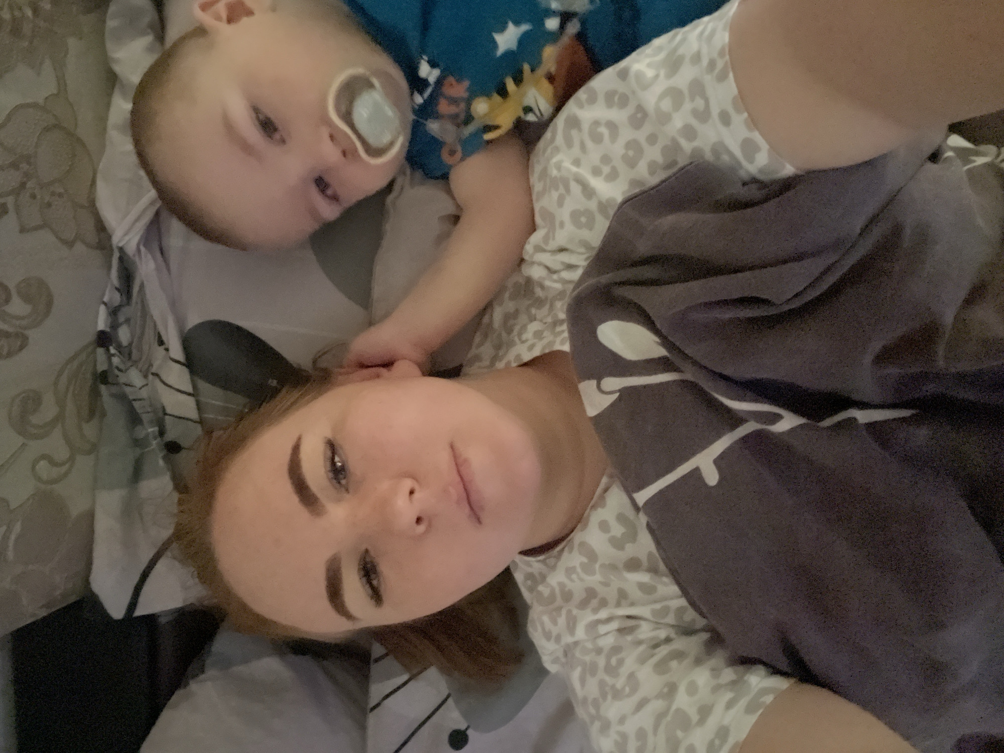 A woman and a baby lie on a patterned gray bed. The baby, dressed in a blue and brown patterned shirt, has a pacifier in its mouth. The woman wears a gray and white patterned shirt. A dark gray blanket covers part of her body. They appear to be indoors. The specific location in Chaikovsky, Russia, cannot be determined from the image.