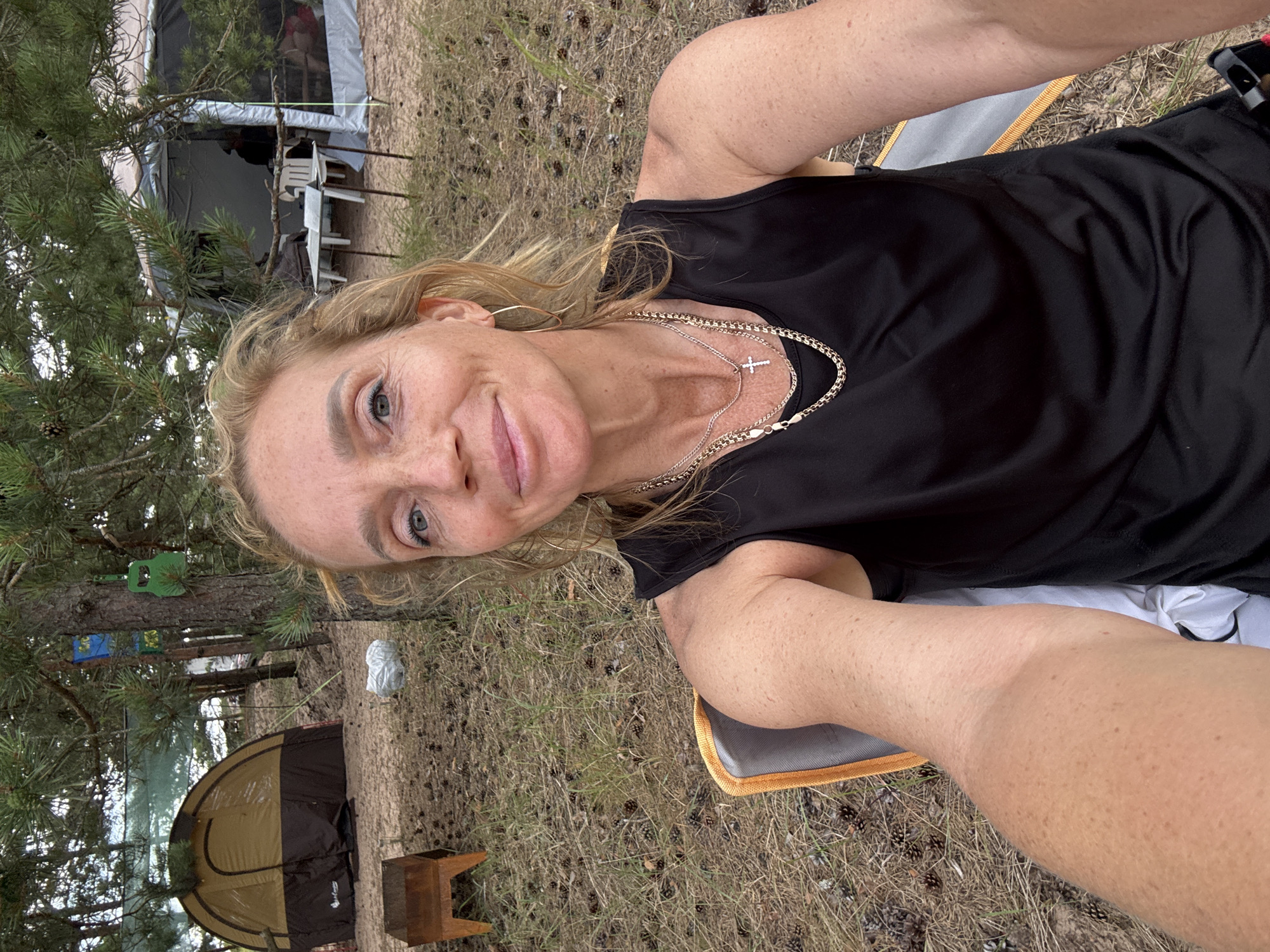 A Caucasian woman with blonde hair and fair skin, wearing a black tank top and two necklaces, one with a cross pendant, sits in a camping chair outdoors. She is looking at the camera with a slight smile. The background shows a campsite with two tents, a portable grill, and trees. The ground is covered in dry grass and pine cones.