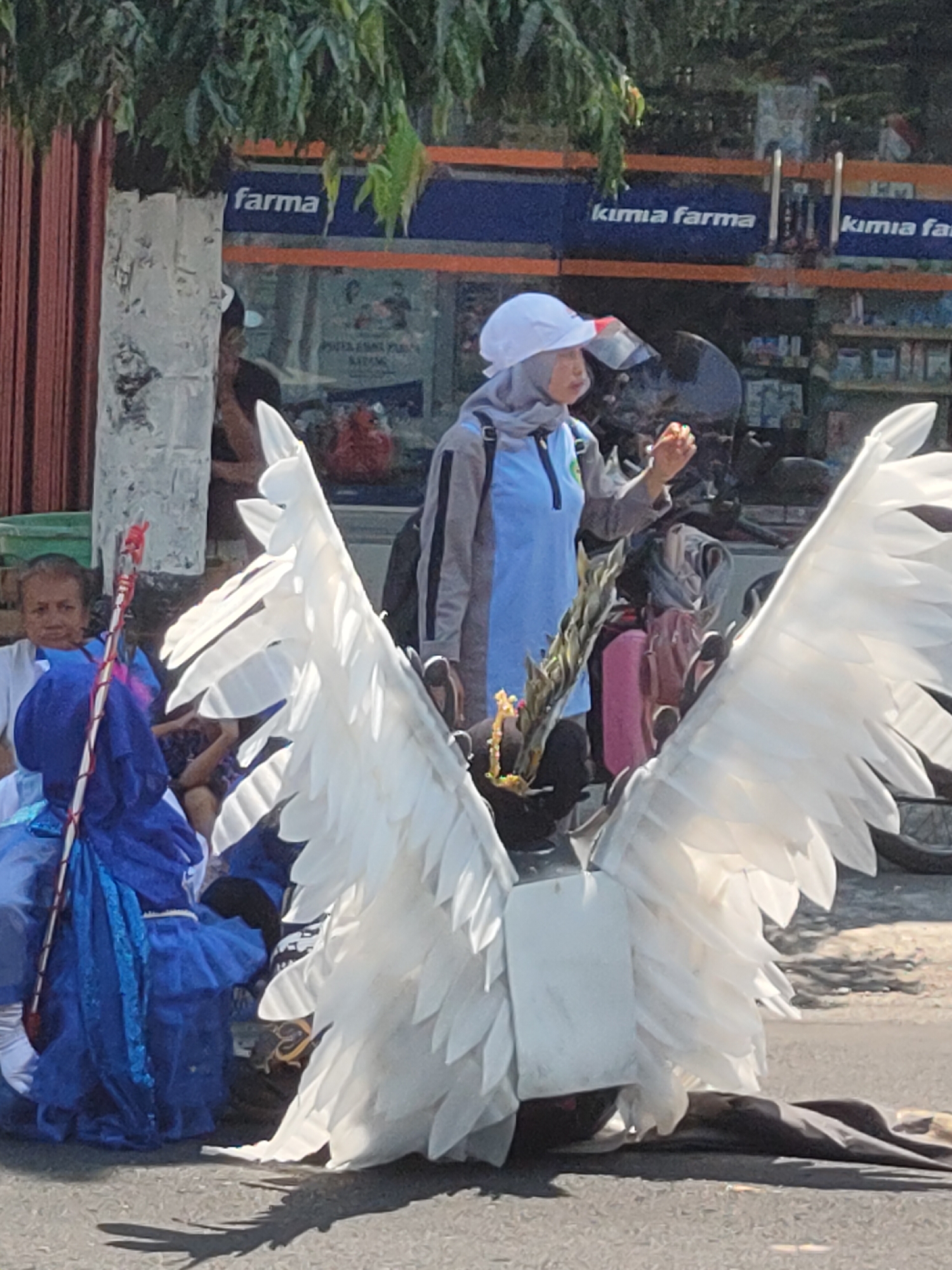 In Ngasem, Indonesia, a person in a large, white feathered costume resembling wings is standing on a street, facing away from the camera. The costume appears to be a prop for a performance or parade. Behind this person, a woman wearing a blue outfit and a headscarf is holding a long, decorated staff. Another woman, partially visible, is seated in the background, wearing a blue garment. In the distance, a storefront with the sign "kimia farma" is visible, suggesting a commercial area. The scene is outdoors, with trees overhead and a paved street.