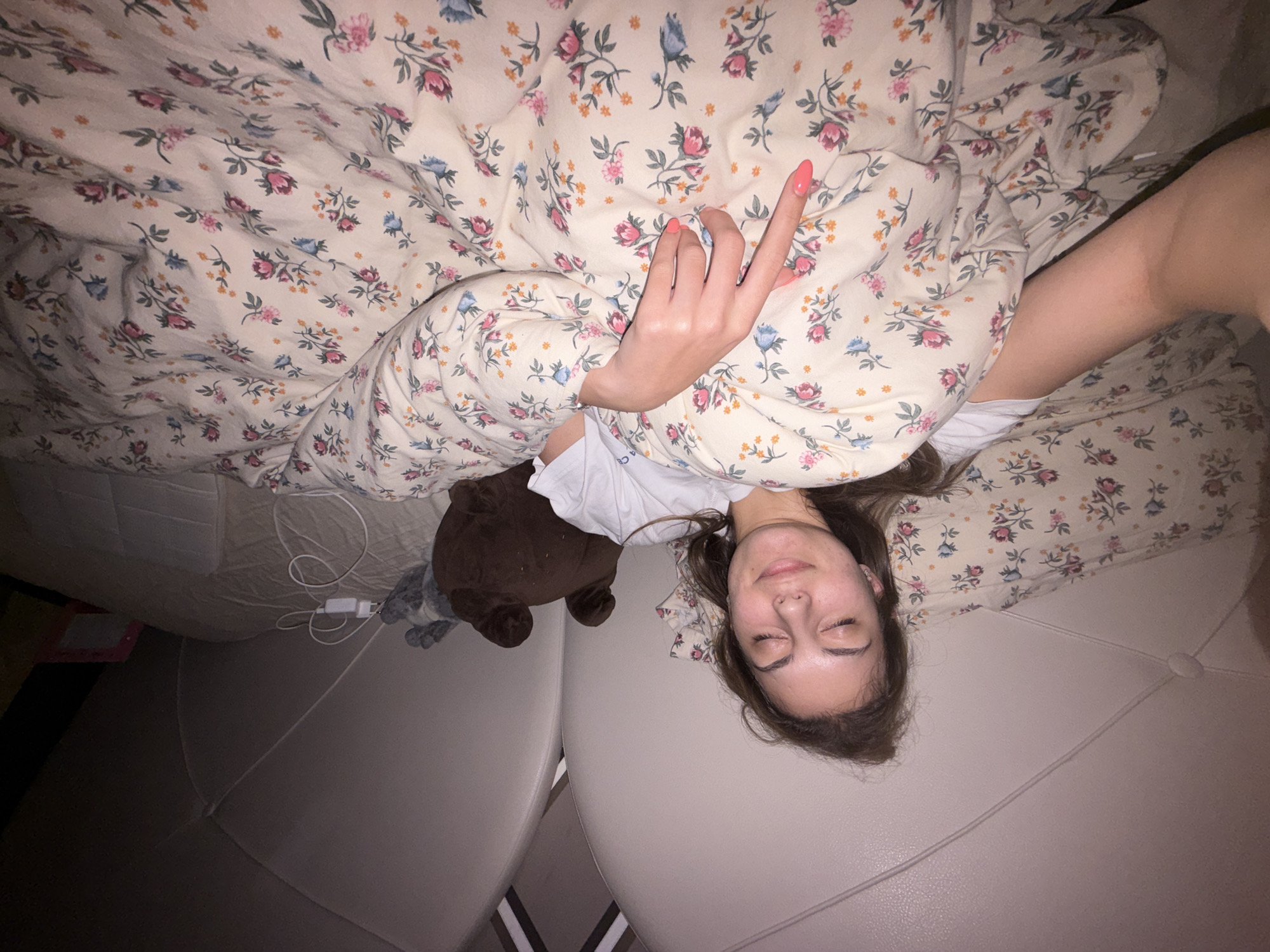 A young woman is lying on a bed with a floral patterned duvet covering her. She has her eyes closed and her hand is visible above the duvet, extended with her middle finger raised. Next to her is a brown teddy bear. A phone charger is lying on the bed. The environment appears to be a bedroom with a light-colored headboard. There is no information about the location in Burgas, Bulgaria.