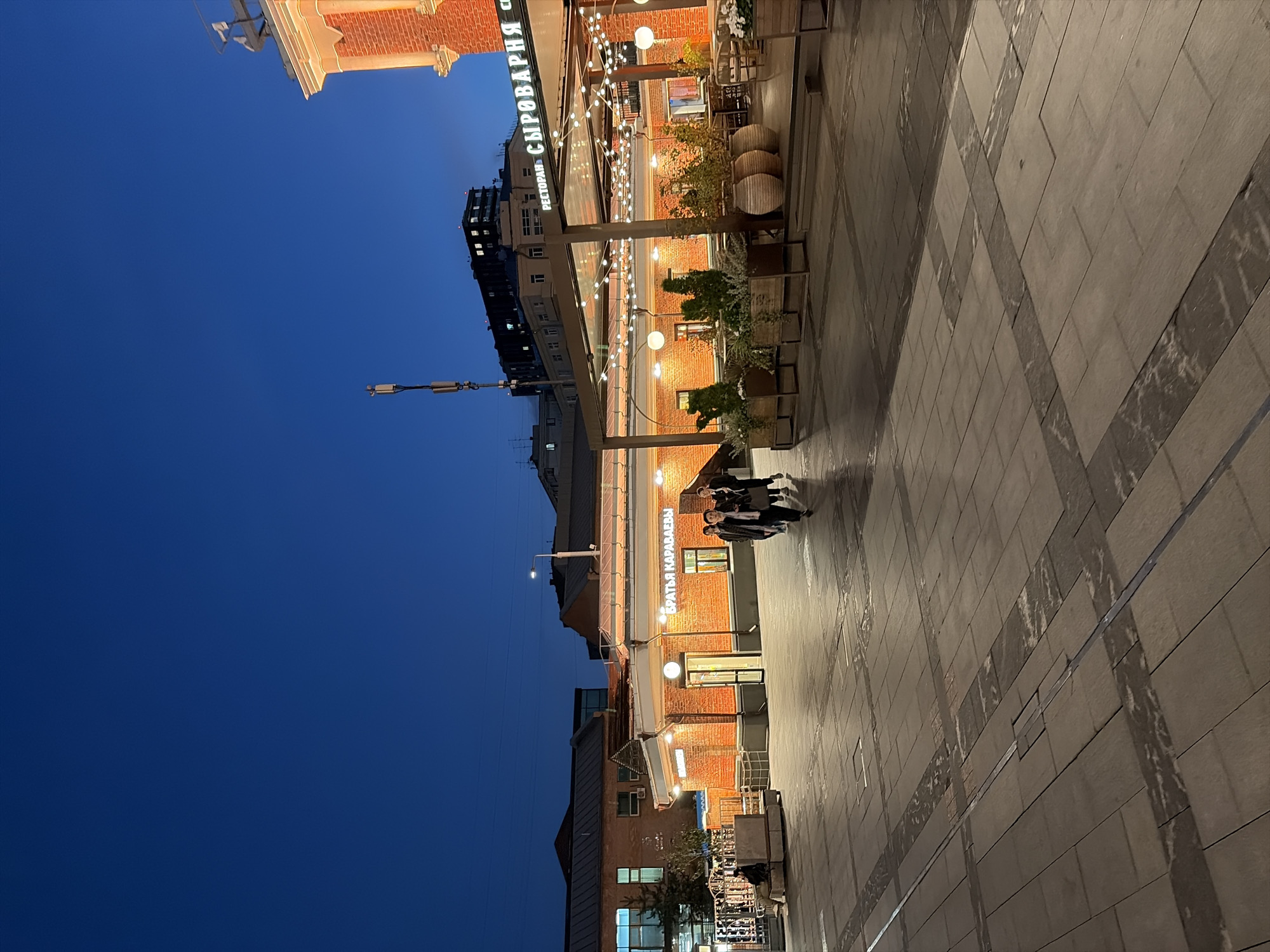 The image captures an outdoor plaza area at dusk in Moscow, Russia, specifically at the Brothers Karavaevy (Братья Караваевы) establishment, indicated by the signage. The scene depicts a pedestrian area paved with large, light-colored rectangular tiles, interspersed with darker stone strips forming a pattern. To the right, a brick building with illuminated windows and a red brick facade is visible, adorned with string lights and large planters containing greenery. This section appears to be an outdoor seating area for a restaurant or cafe. In the mid-ground, a lower brick building with arched openings and signs is present, with several people walking in front of it. The sky above is a deep, clear blue, transitioning from twilight to night. Further in the background, modern apartment buildings and a cell tower are visible against the darkening sky. One person is seated on a bench on the left side of the image, facing away from the camera. The overall impression is one of a lively urban evening setting.