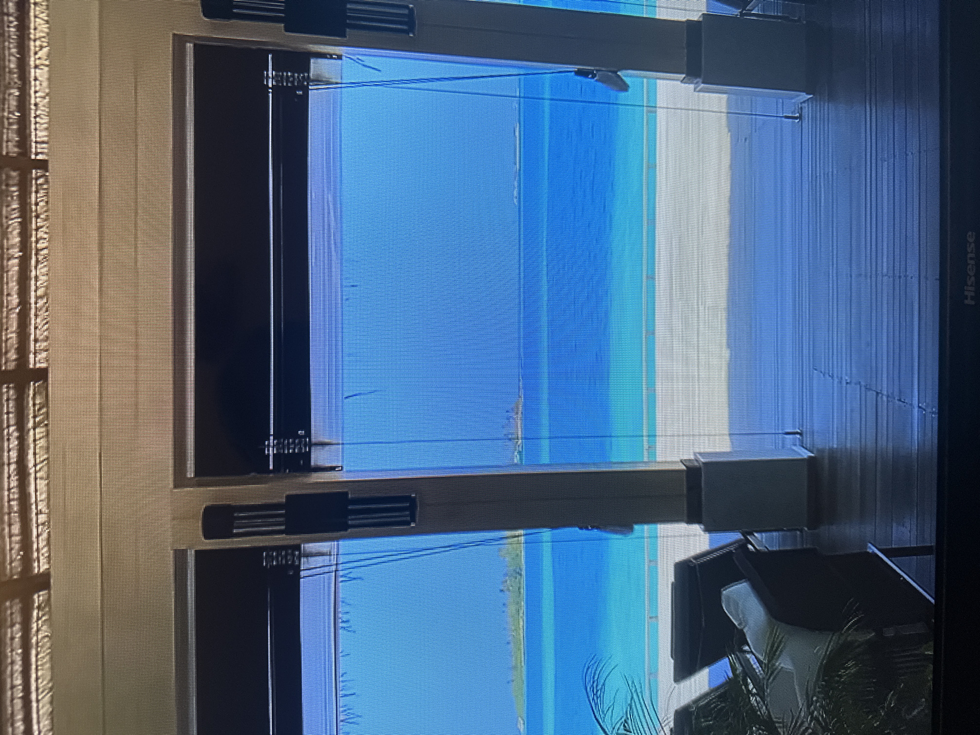The image depicts a view through an open doorway, looking out onto a tropical ocean scene. The doorway is framed by wooden posts and has dark blinds pulled down on either side. Beyond the doorway, there is a clear, vibrant blue ocean, with a narrow strip of white sand and a line of palm trees visible on a distant island. In the foreground, on a wooden deck, are two dark lounge chairs with white cushions, and a large palm frond. The lighting suggests it is daytime, and the overall atmosphere is serene and inviting. The image does not contain any elements that would indicate it is located in Kyiv, Ukraine.