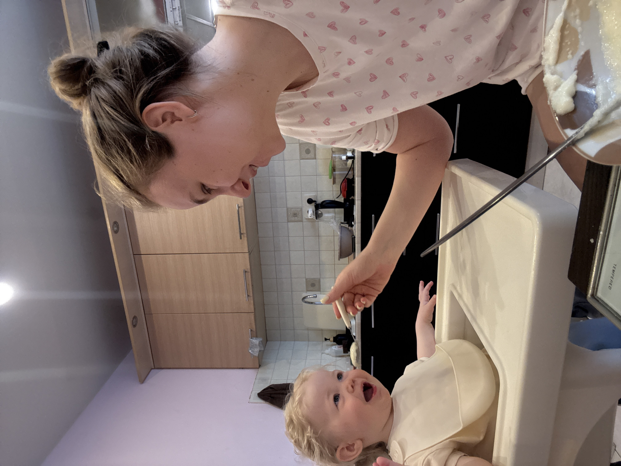 A woman in a white shirt with pink hearts feeds a baby in a high chair. The woman holds a spoon with food in a clear bowl. The baby is wearing a beige bib. The scene appears to be indoors, possibly in a kitchen, with cabinets and tile visible in the background. The location is Unknown, Unknown.