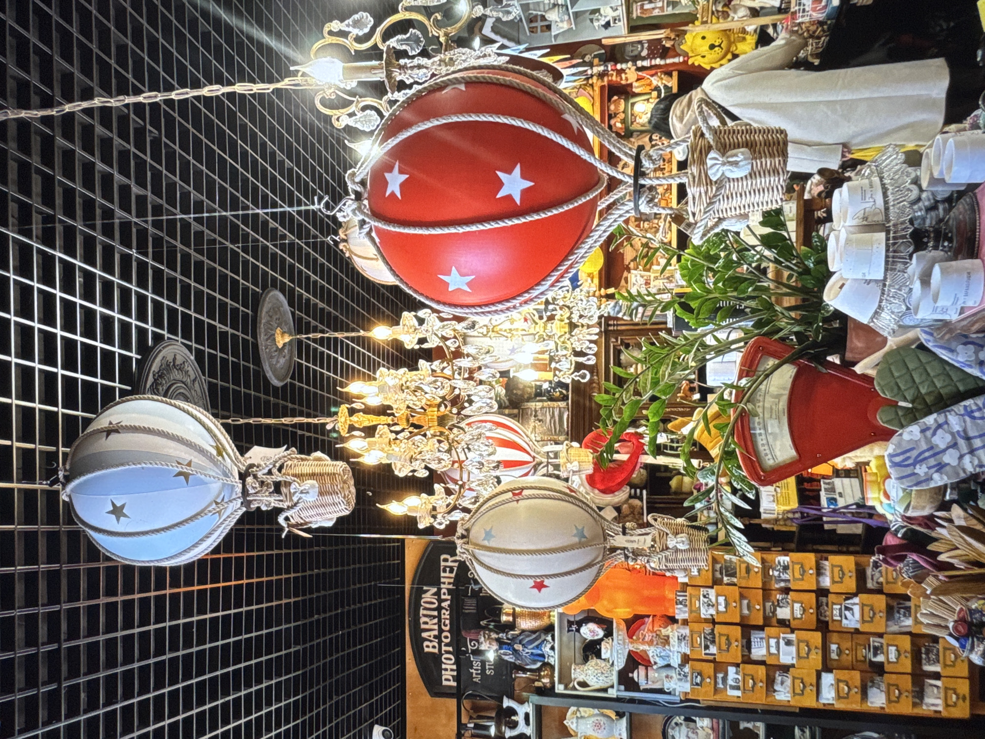 The image is a medium shot, taken from a slightly elevated perspective, of a whimsical shop interior. The scene is dominated by several decorative hot air balloon models suspended from the ceiling, each with a woven basket attached. One balloon is bright red with white stars, another is pale blue and white with gold stars, and a third is white with red stars. The balloons are illuminated by a golden chandelier with crystal accents, casting a warm glow.
Behind the balloons, the shop is filled with various items, suggesting a vintage or antique store. Shelves are packed with a collection of objects, including numerous small drawers or boxes, some displaying photographs or postcards. Other visible items include a red vintage scale, a green oven mitt, a white coat, and a woven basket with a bow. Lush green foliage spills from a pot, adding a natural element to the scene. The overall impression is cluttered yet charming, evoking a sense of discovery and nostalgia.
The image appears to be taken indoors, likely during daylight hours given the bright lighting, though the specific time of day cannot be definitively determined. There are no discernible weather cues. The setting is suggestive of a quaint boutique or a curated marketplace.
Visible text includes "BARTON PHOTOGRAPHER ARTIST STUDIO" on a black sign. The location context provided is Novinki, Russia.
