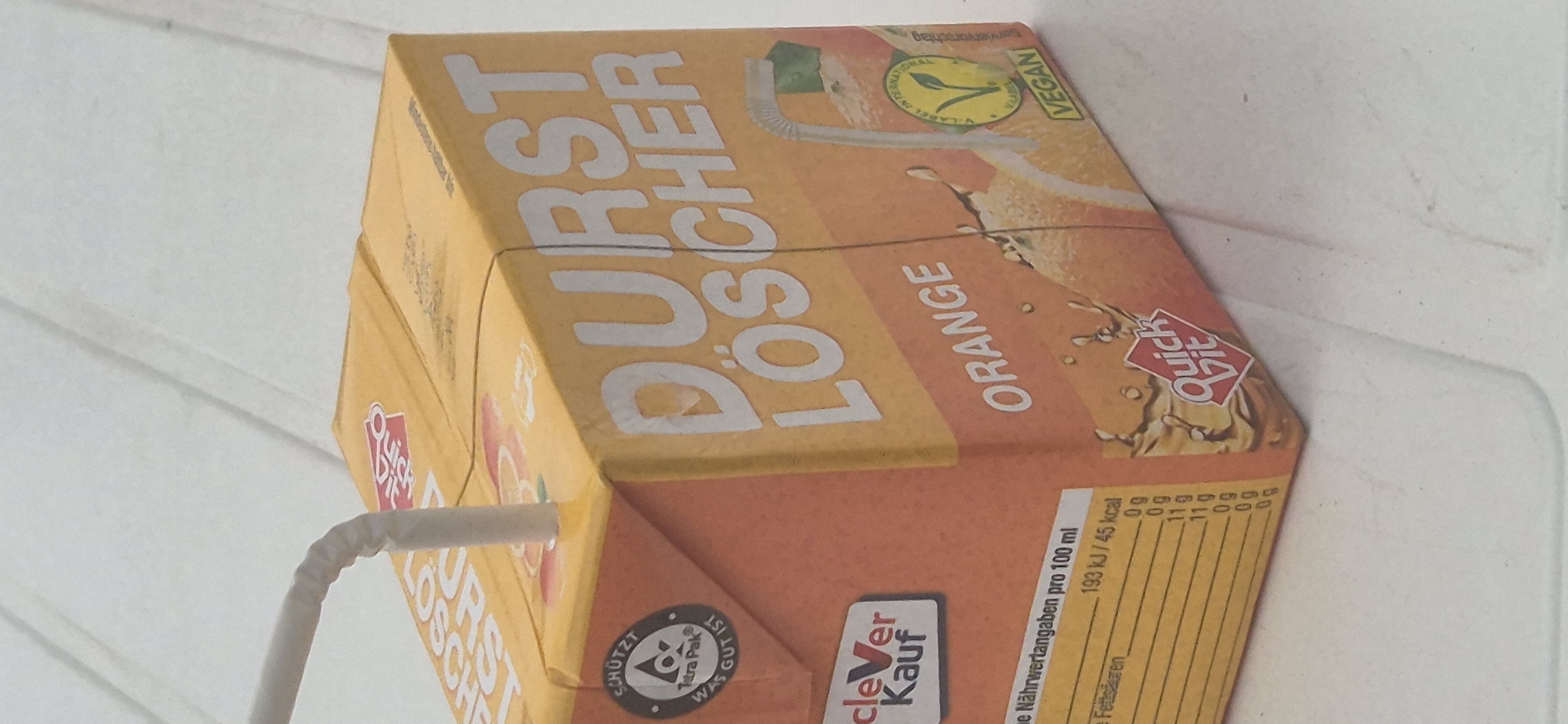 A Tetra Pak juice carton, labeled "DURST LÖSCHER ORANGE," is lying on a white, textured surface that resembles a table. The carton is orange and yellow and features a straw inserted through the top. A nutrition label and the "cleVer Kauf" logo are visible on the side of the carton. There is no indication of the city of Mainz, Germany, in the image.