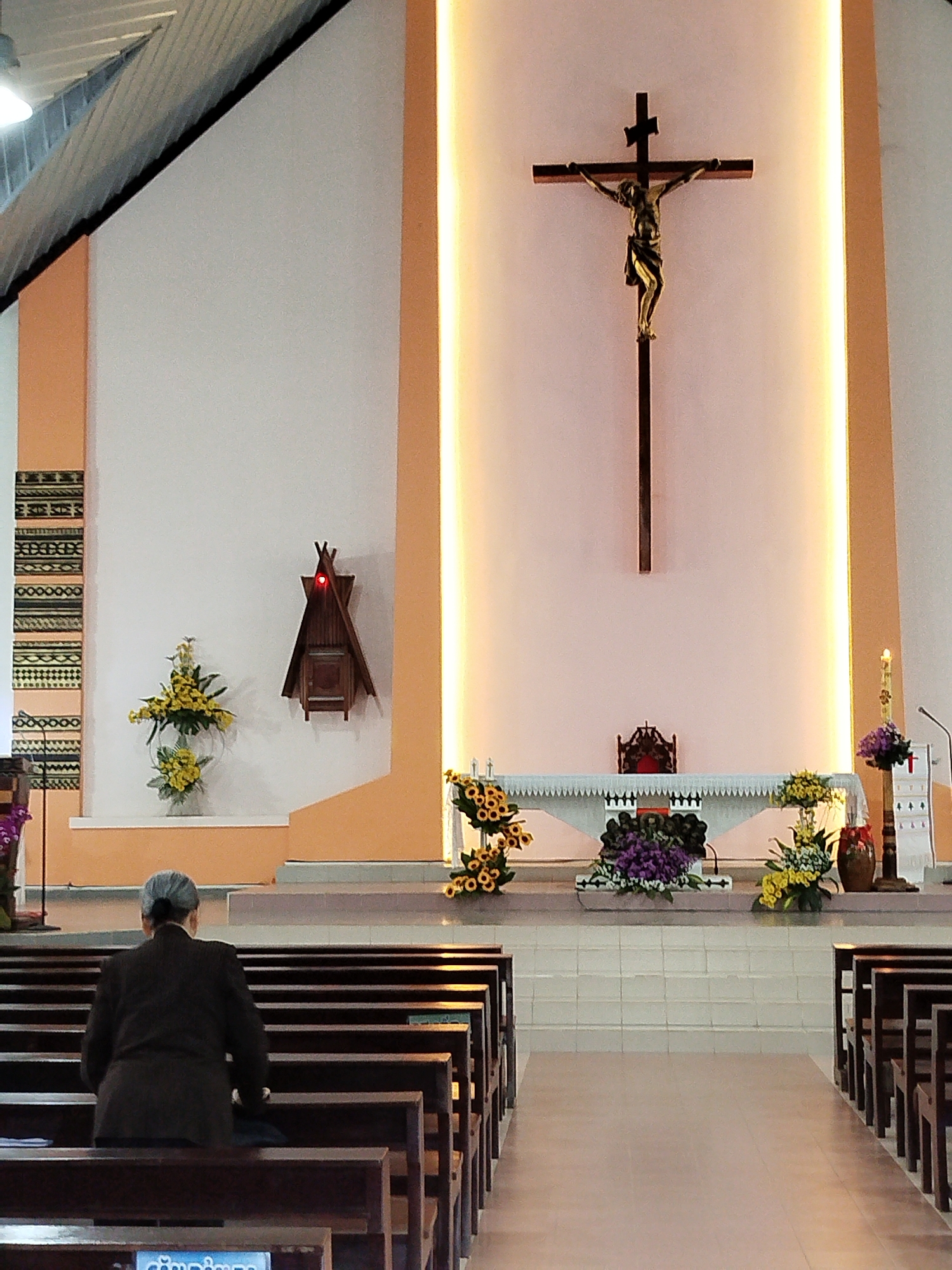 A view from inside a church located in K’Long, Vietnam. The church features a central altar with a large crucifix mounted against a softly illuminated panel. The altar is adorned with various floral arrangements. In the foreground, a woman with grey hair is seated in a pew, facing the altar. Wooden pews line the aisle, creating a path toward the altar. The walls are painted in light shades, and cultural artifacts are displayed on the left side of the altar area. The architecture of the church is simple, with a modern design, and the overall setting suggests a peaceful, contemplative atmosphere.